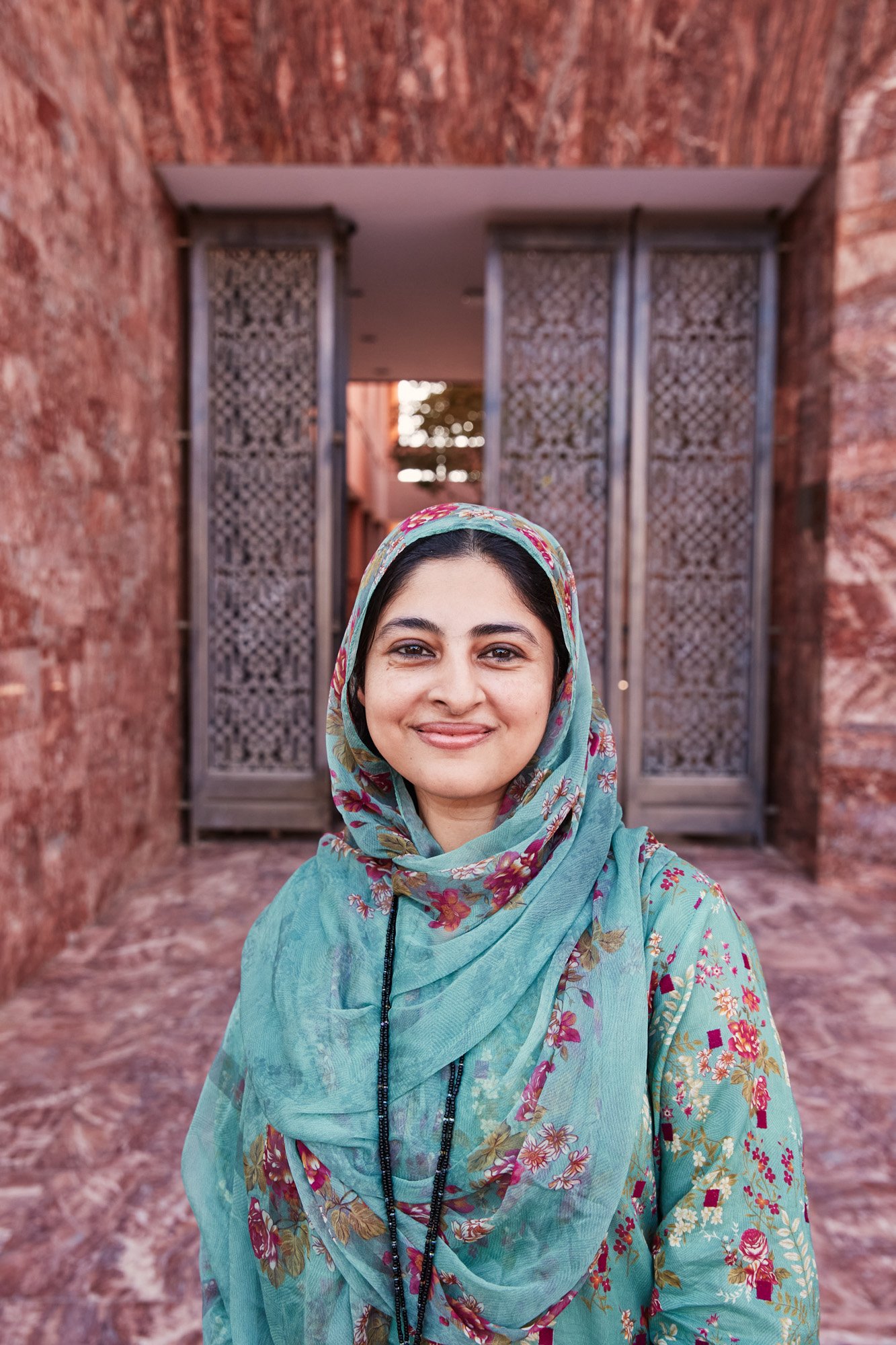 A woman wearing a floral headscarf and matching floral top standing in front of a red stone wall with wooden lattice doors at Aga Khan Hospital in Karachi Pakistan.