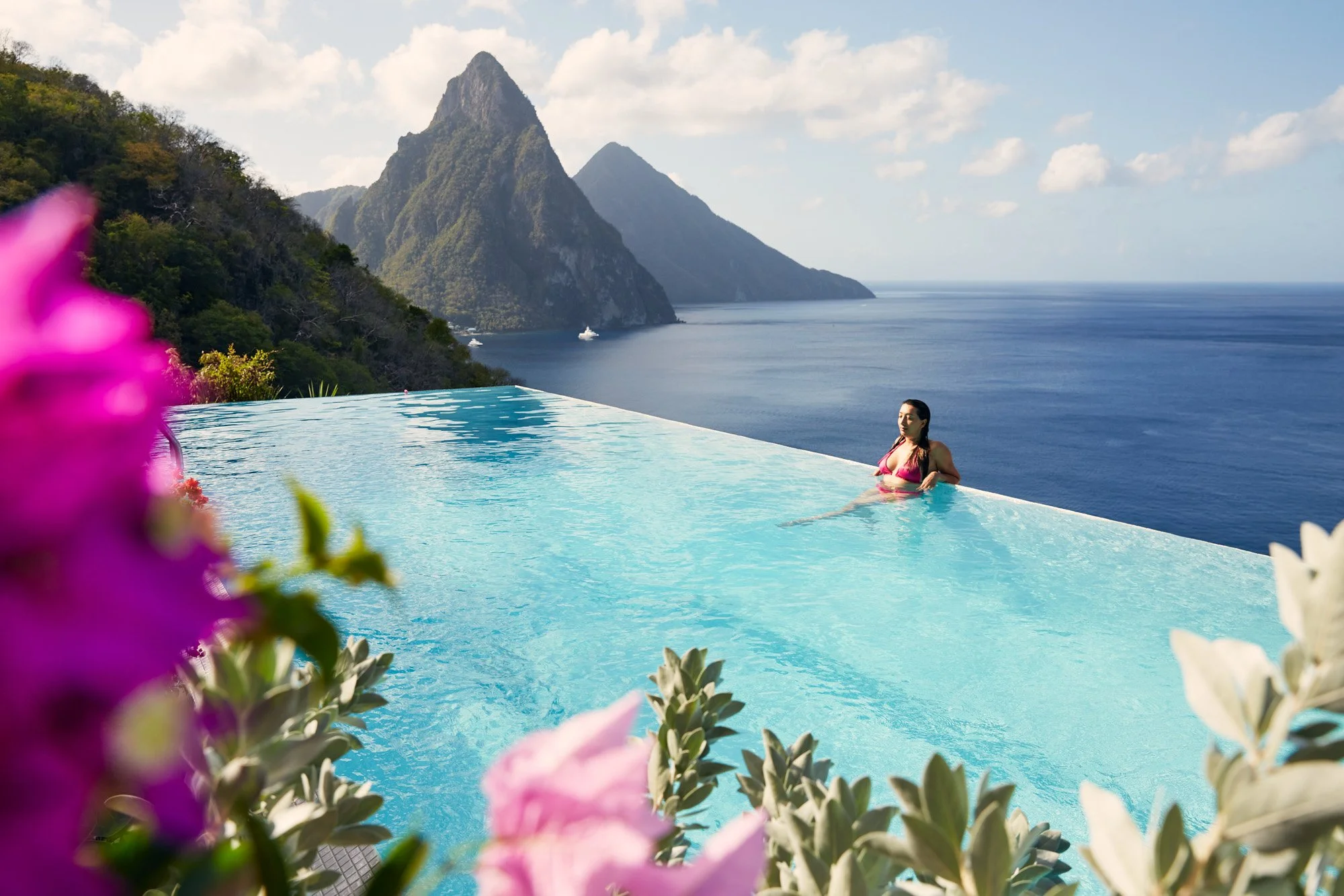 A woman in a pink bikini relaxing in an infinity pool overlooking the ocean in St Lucia with lush green mountains the pitons in the background and colorful flowers in the foreground.