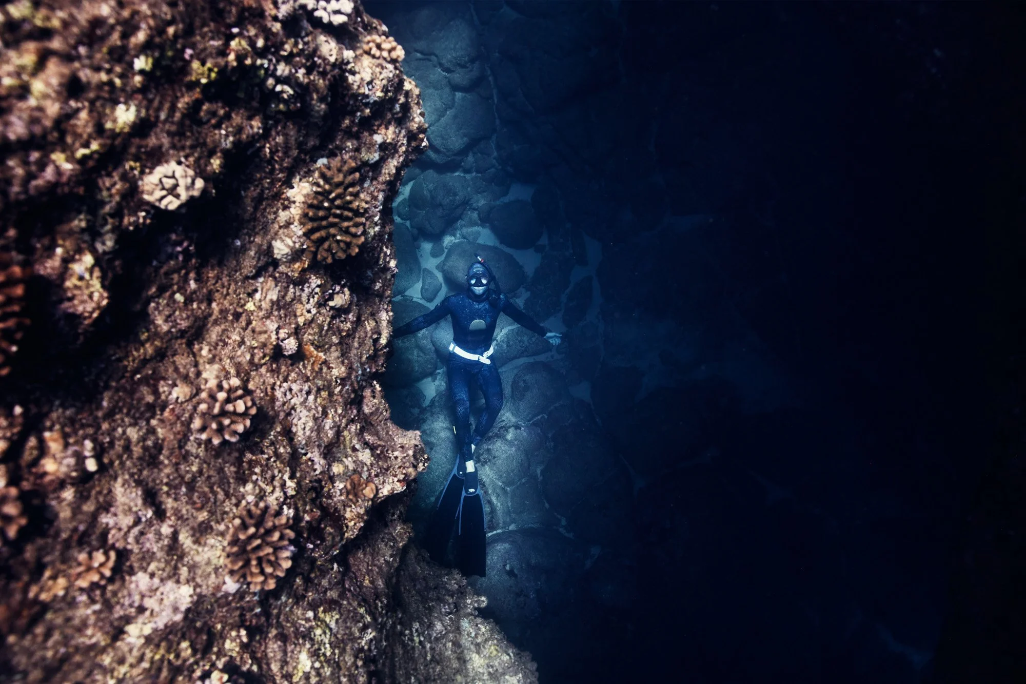 A man in free dive gear laying on the ocean floor, underwater next to a coral reef, smiling, with rocks in the background.