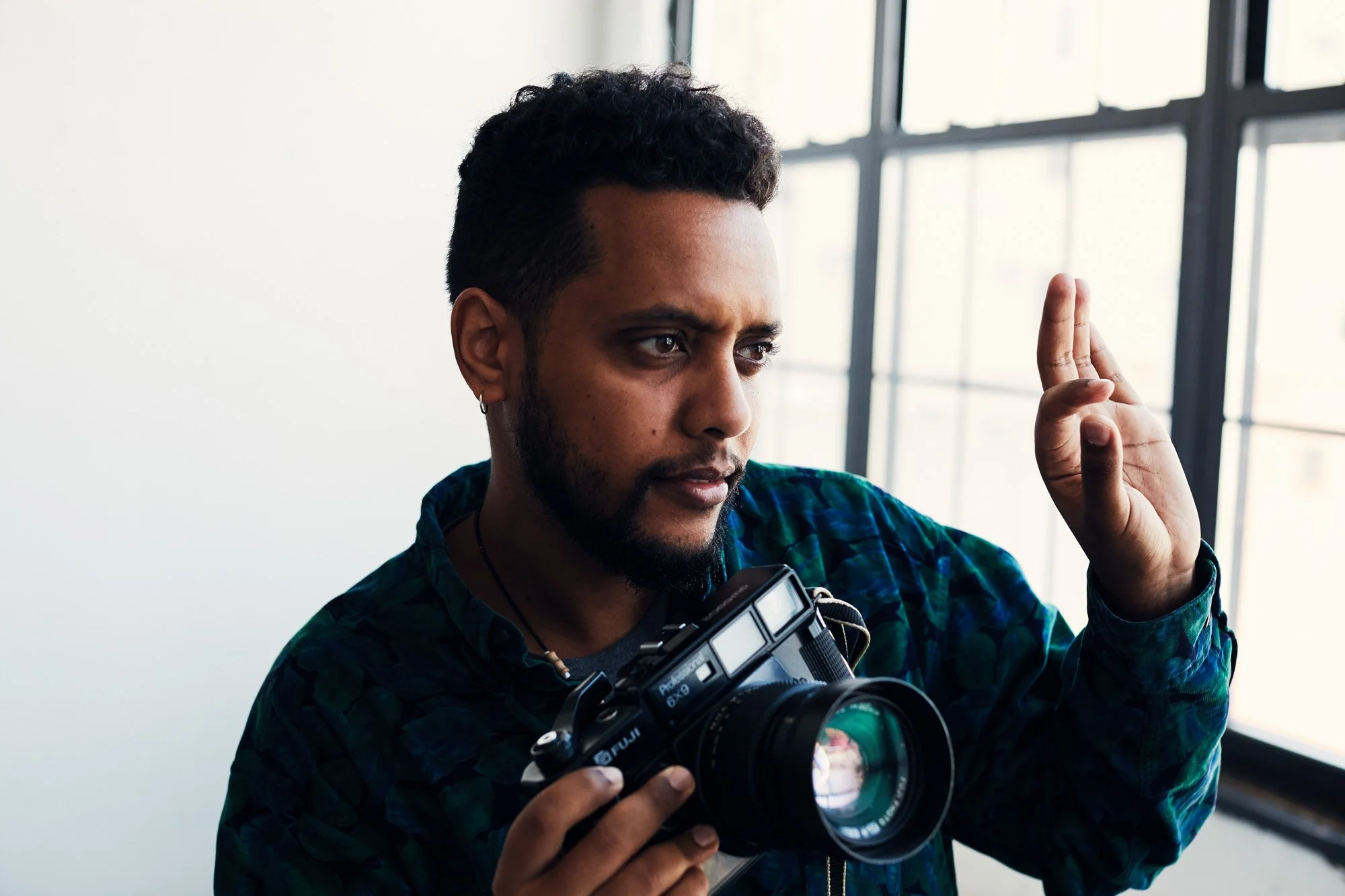 Photographer Mehron Menghistab with dark hair and a beard holding a camera, gesturing with his right hand, standing next to a window.