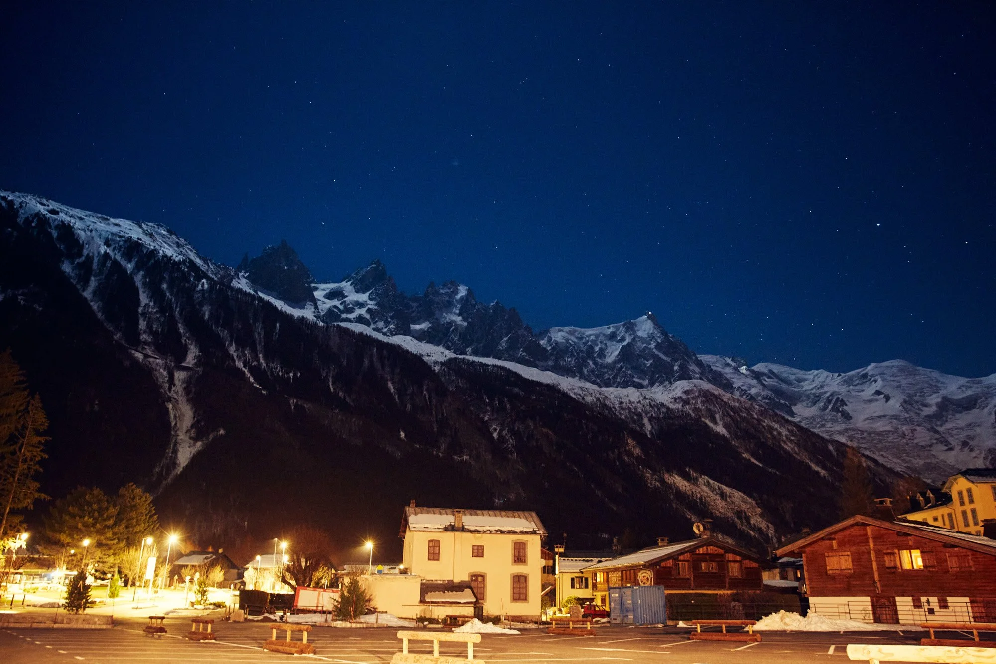 Night view of snow-covered mountains against a starry sky in Chamonix, France, with a small town or village in the foreground, including a parking lot and illuminated buildings.