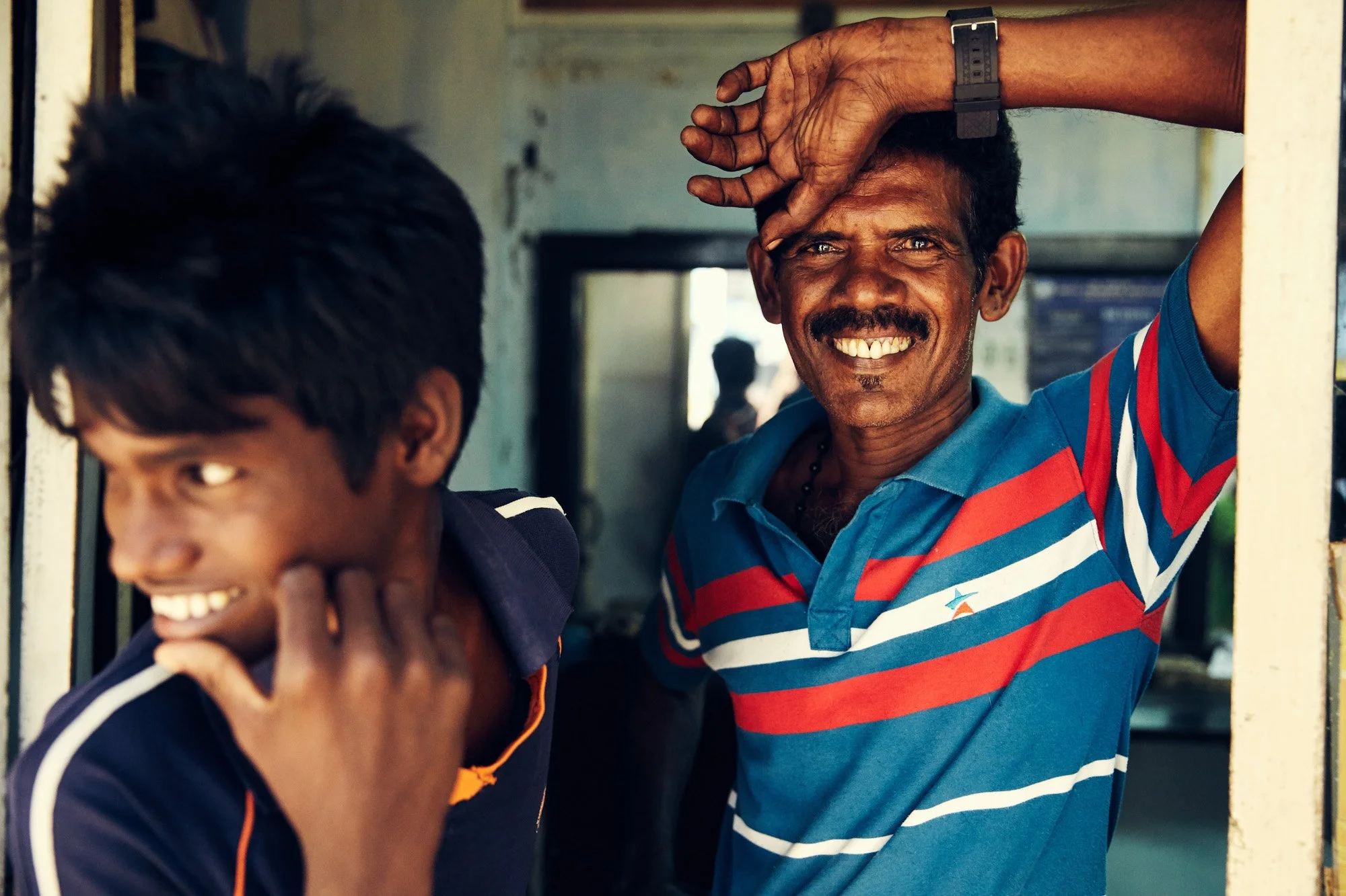 Two men smiling in Jaffna Sri Lanka, one with a striped blue and red shirt and the other with a dark shirt, standing in a doorway, with happy expressions and relaxed body language.