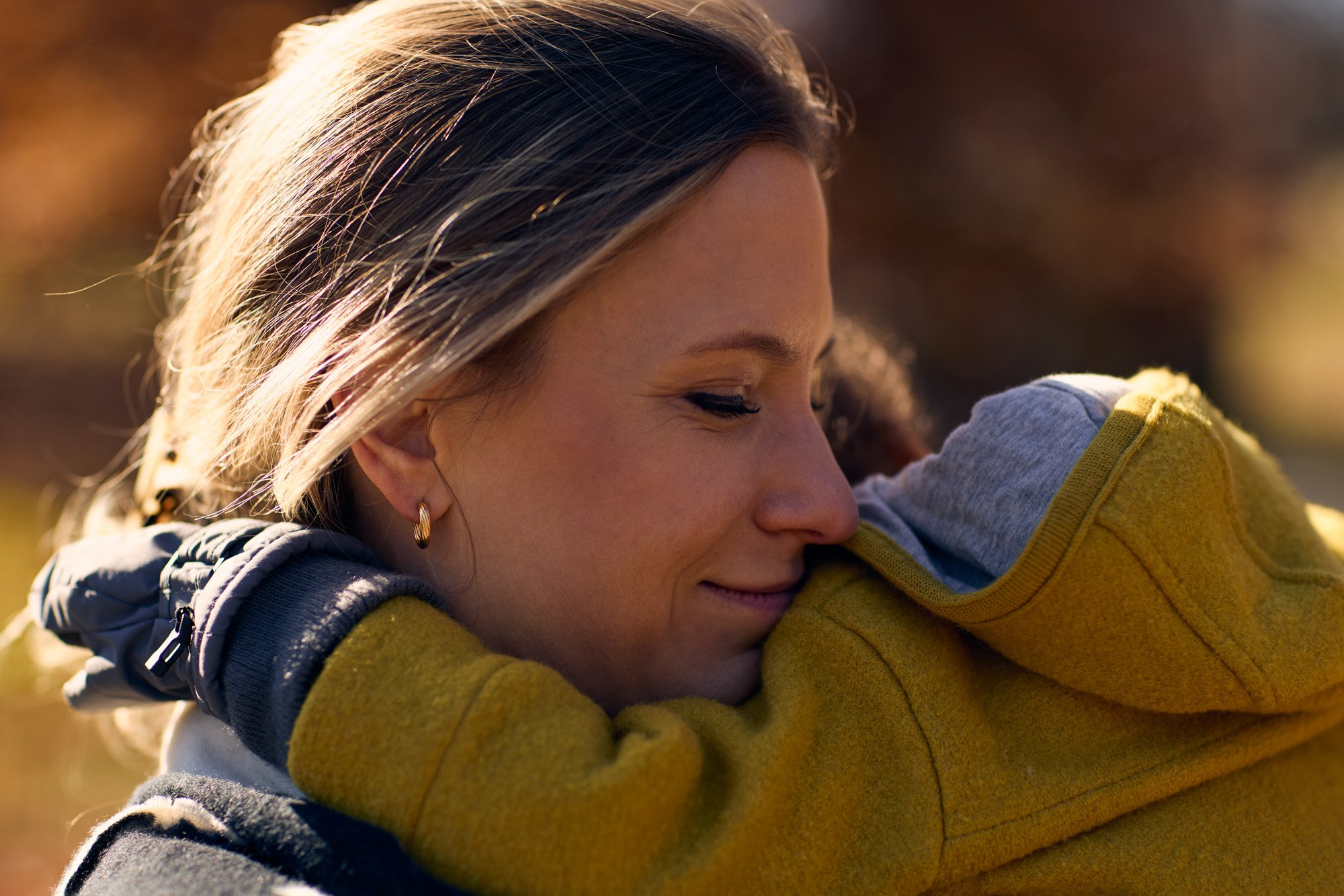 A woman with closed eyes embraces her little boy outdoors, smiling softly, wearing a yellow and gray jacket with a hood, and has earrings fitting the fall season.