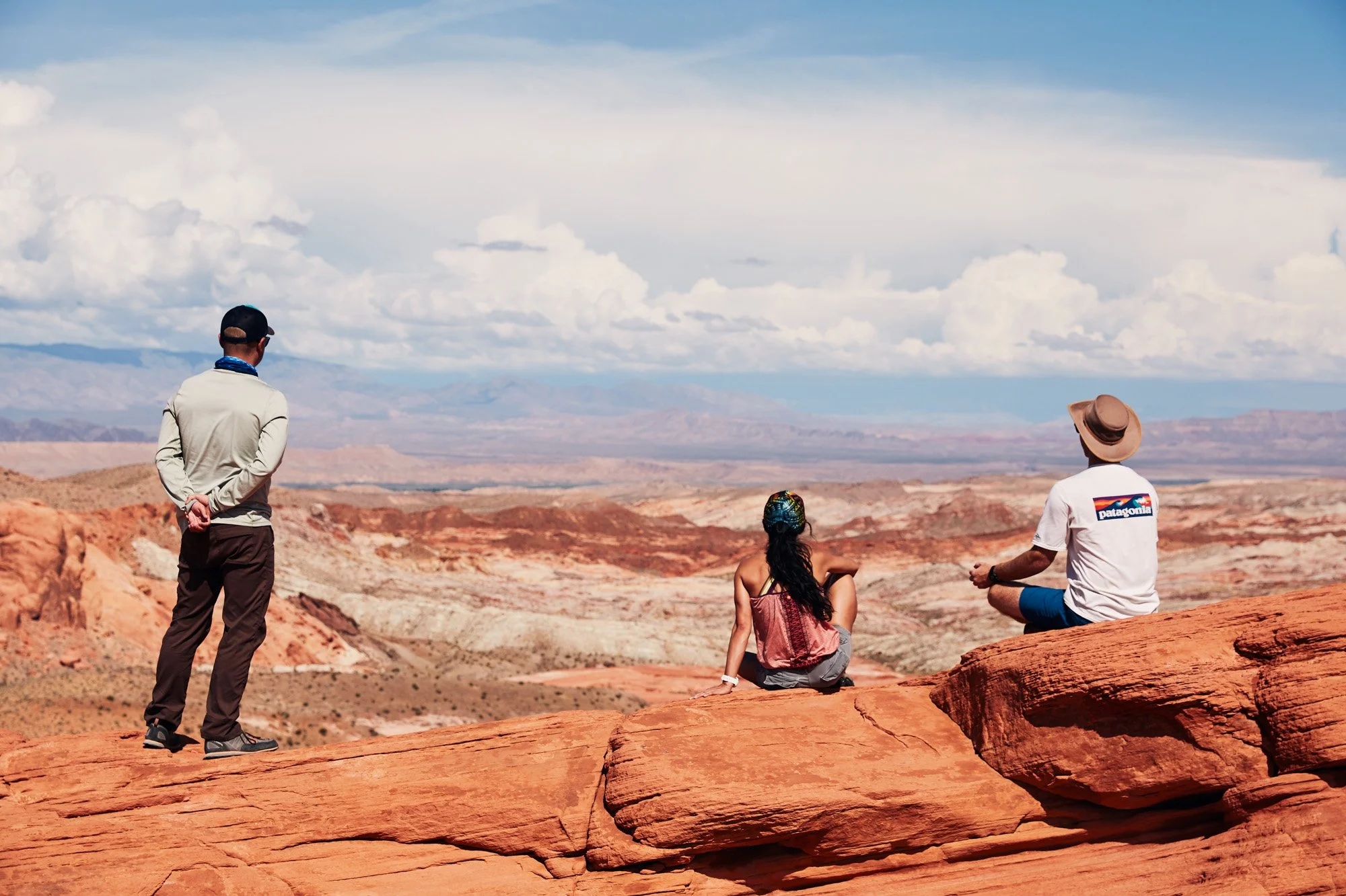 Three people enjoying a desert landscape view in the Valley of Fire Nevada, with two sitting and one standing on red rocks, under a blue sky with clouds.