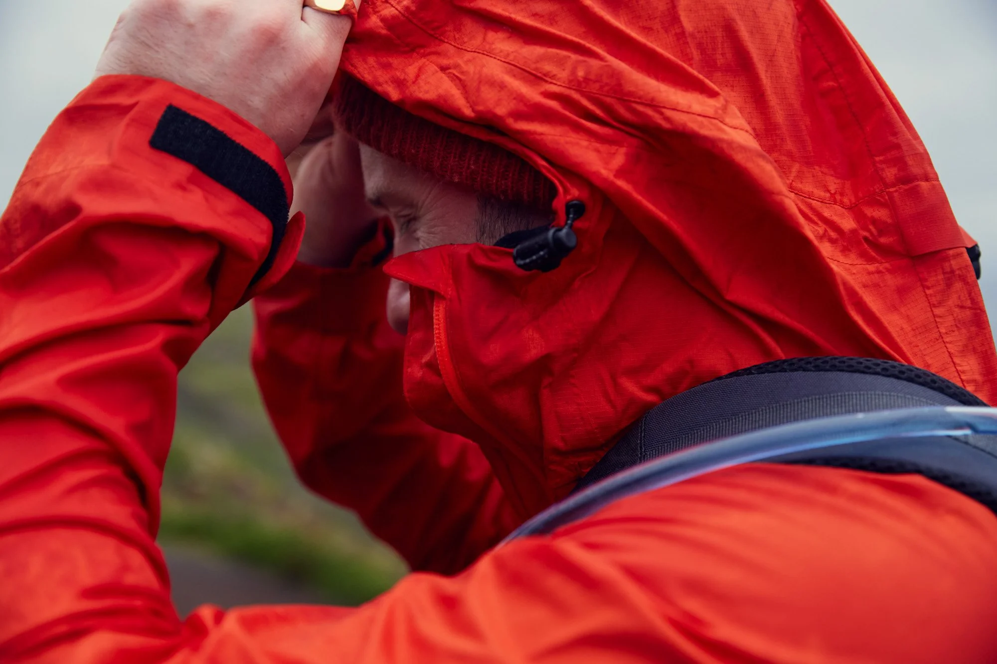 Person wearing a red jacket and hat, adjusting hood outdoors, with a blurred landscape background.