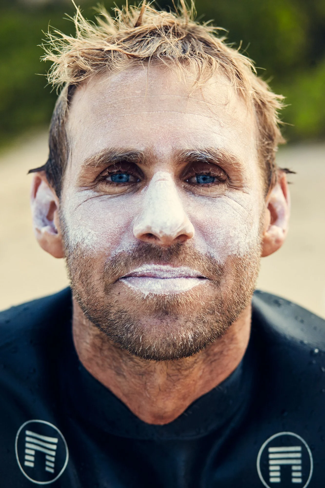 Close-up of big wave surfer Aaron Ungerleider, with blue eyes and light brown hair, wearing a black wetsuit, covered in white foamy soap or sunscreen, outdoors with a blurred green background.
