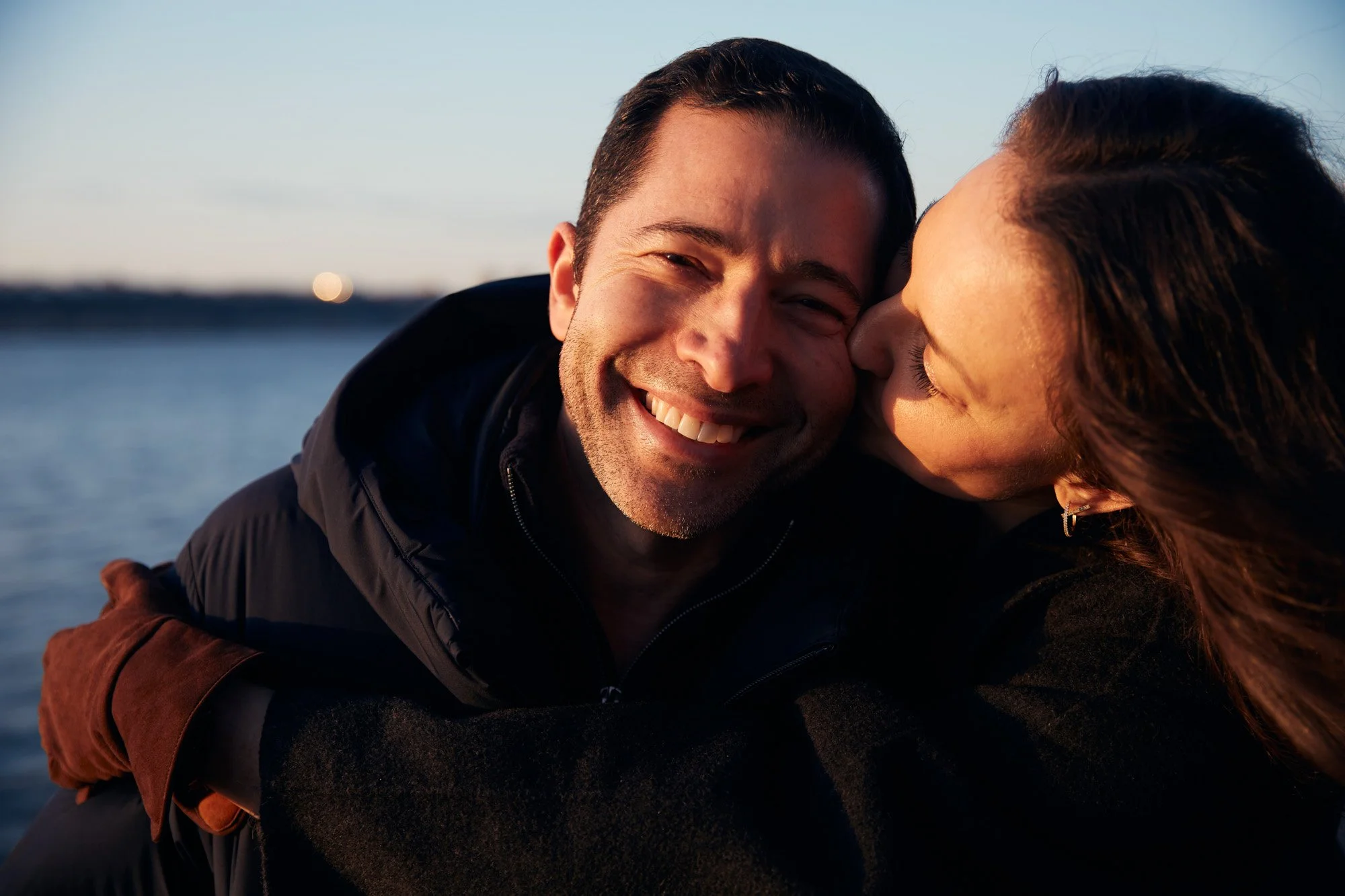 A man and woman sharing a joyful moment near water at sunset, with the woman kissing the man on the cheek.