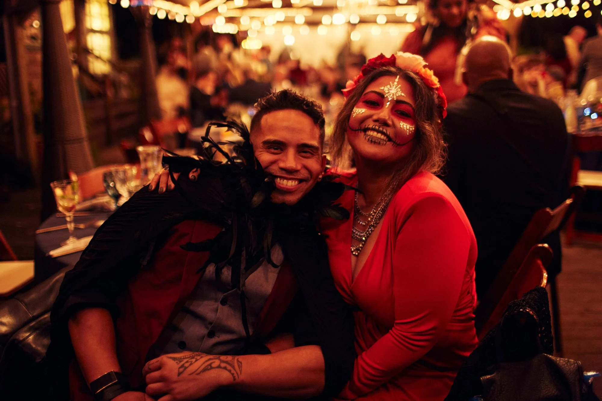 Two people smiling and dressed festively at a lively party or celebration, with colorful face paint and costumes, surrounded by other guests and warm lighting.