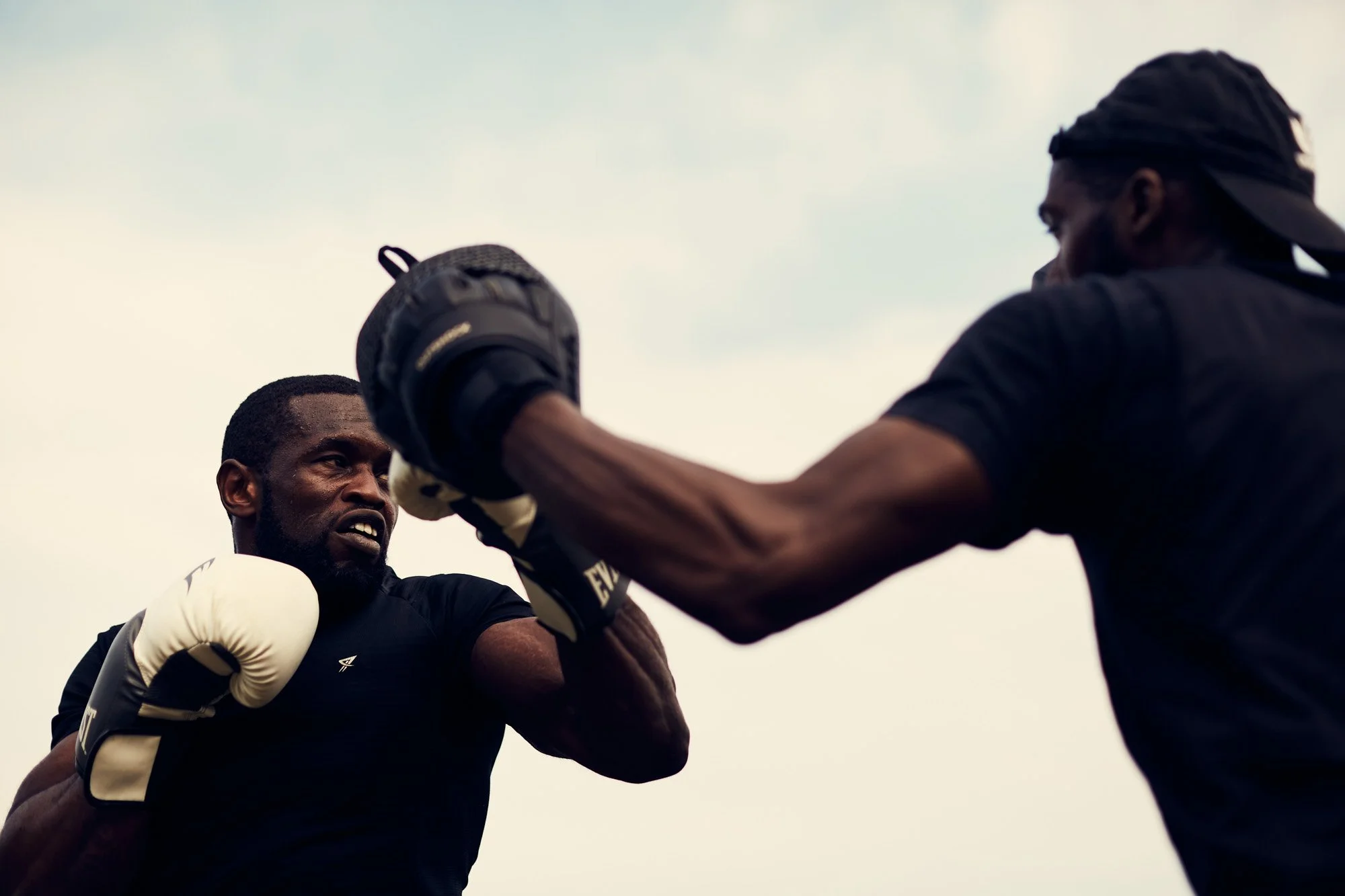 Two Black men training with boxing gloves outdoors, one throwing a punch at the other.