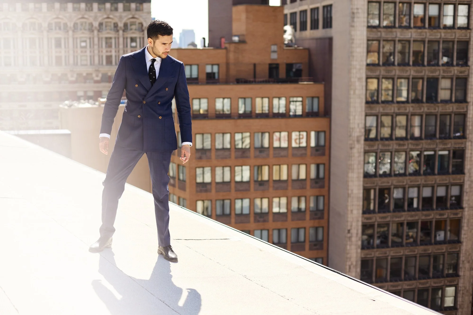 Magician Dan White in a navy double-breasted suit and tie standing on a white rooftop, looking downward, with New York City in the background.