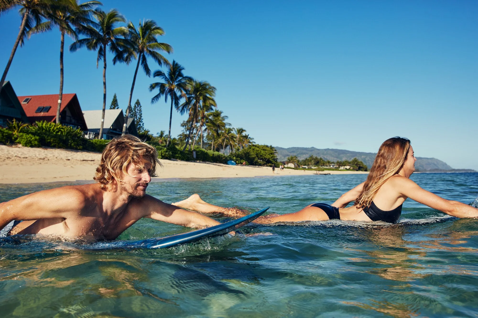 A man and woman are paddling their surfboards in the ocean near a sandy beach with palm trees and houses in the background, enjoying sunny weather on the north shore of Oahu Hawaii
