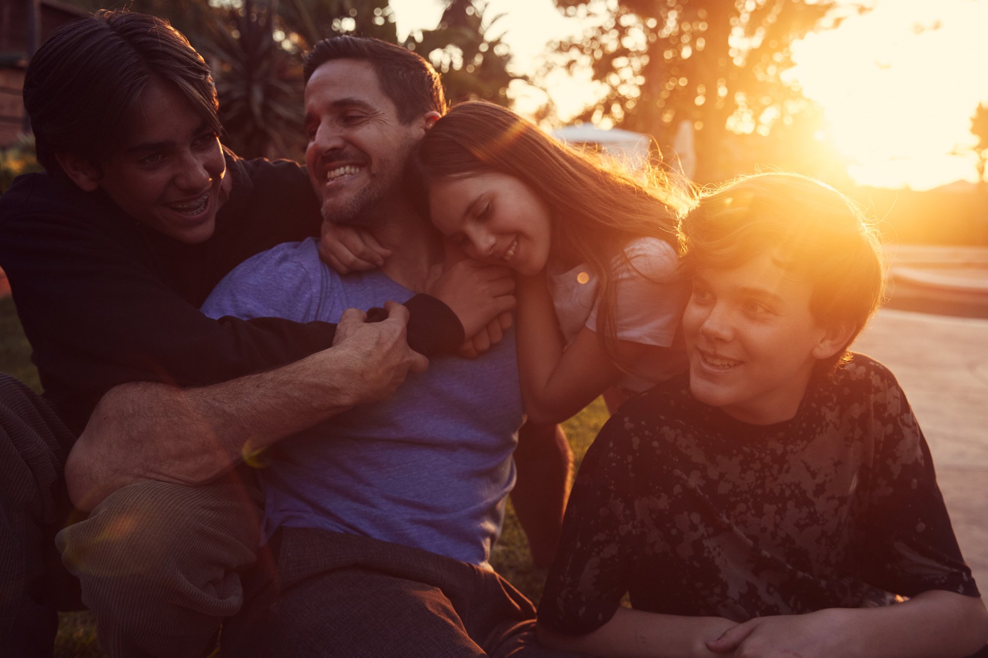 A group of four people, including a father and his children, three younger individuals, smiling and enjoying each other's company outdoors during sunset.