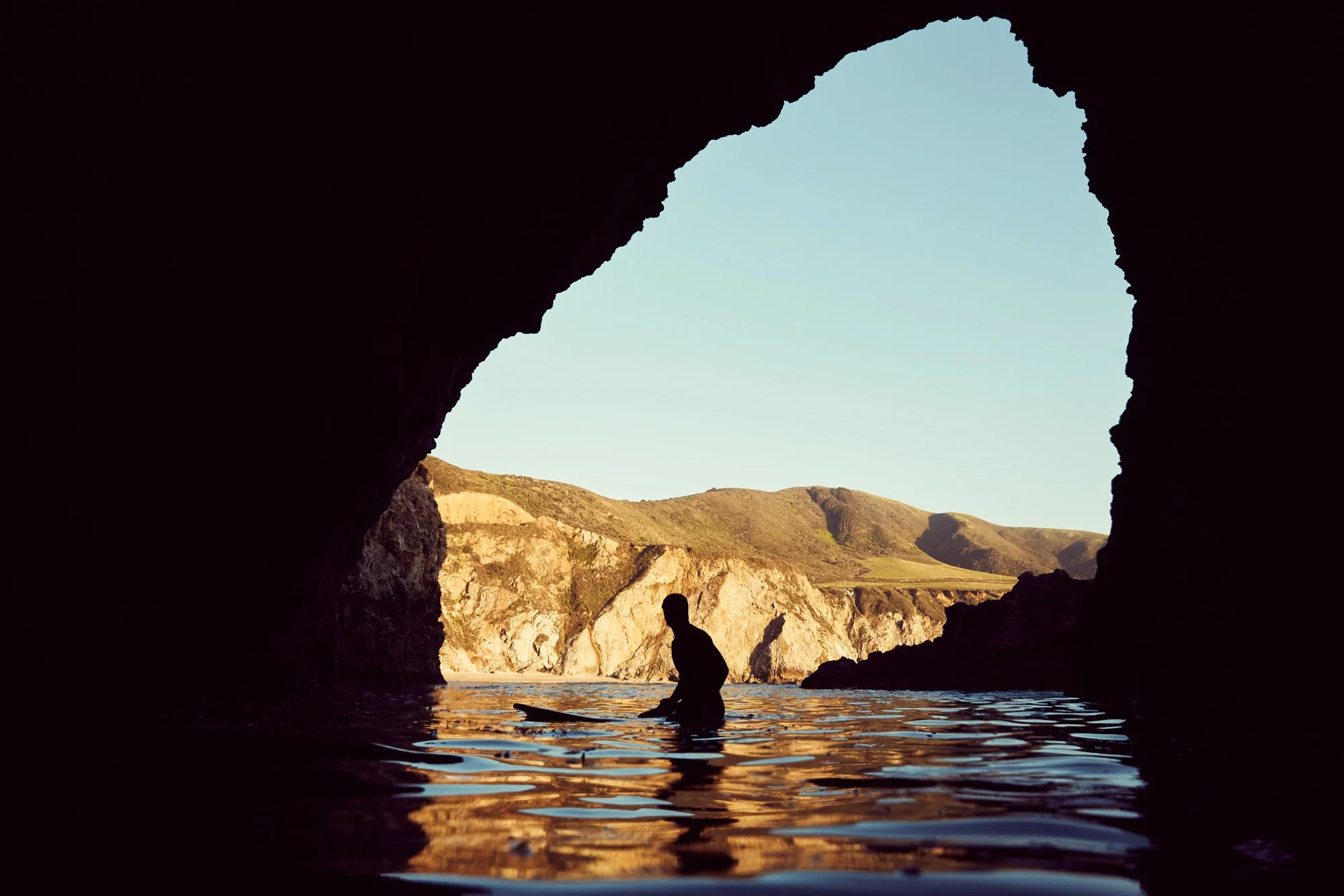 Person sitting on a surf board in a water cave with an opening revealing hills and a clear sky, in Big Sur California