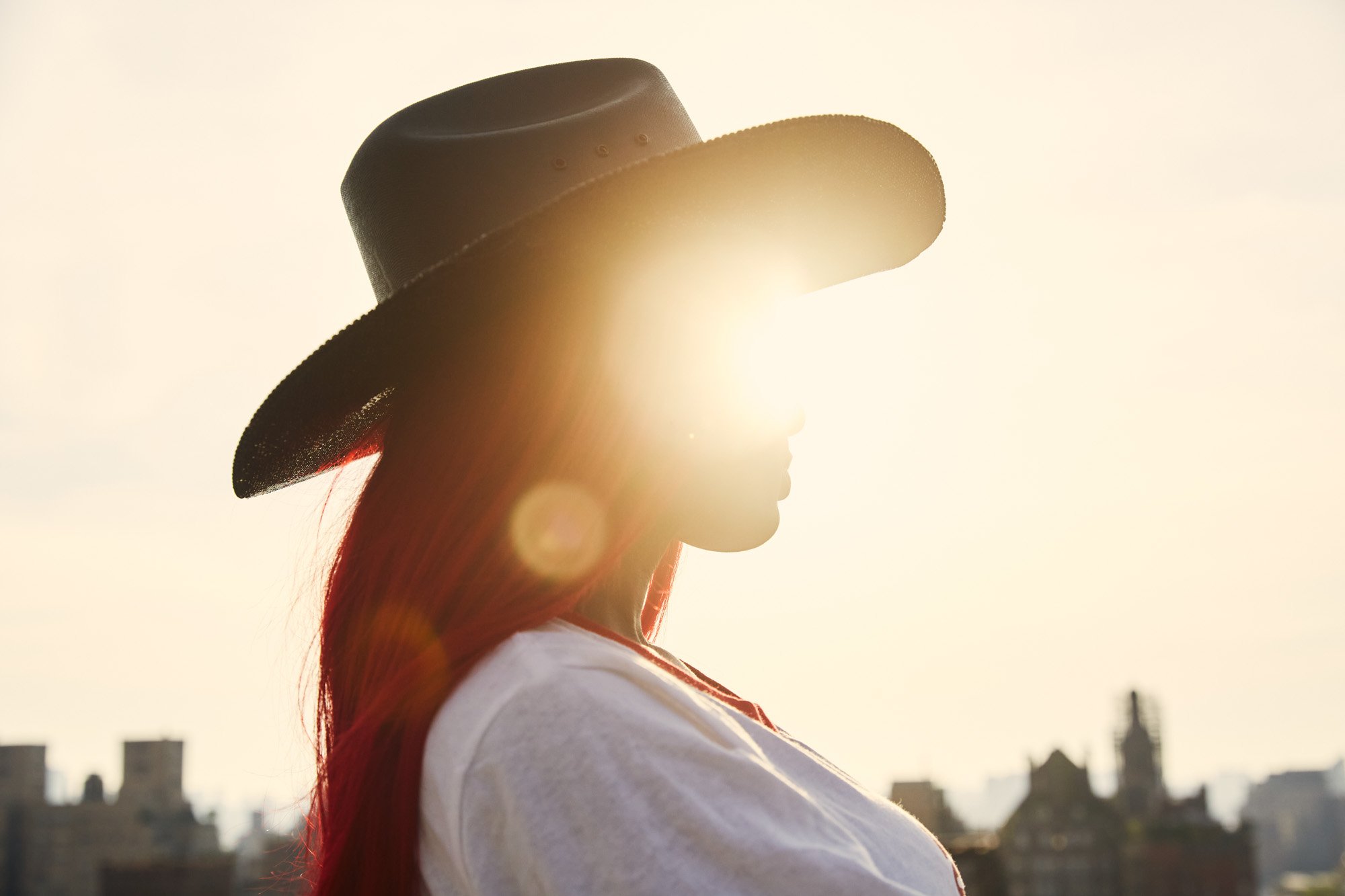 Side profile of singer Reyna Roberts with long red hair wearing a wide-brimmed hat. The sun is shining brightly behind her, creating a backlit silhouette against a cityscape.