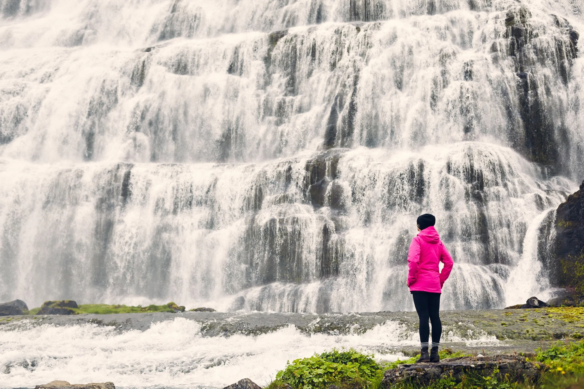 A young woman wearing a pink jacket, black pants, and black boots stands on rocky ground near a waterfall, observing the cascading water in Iceland.