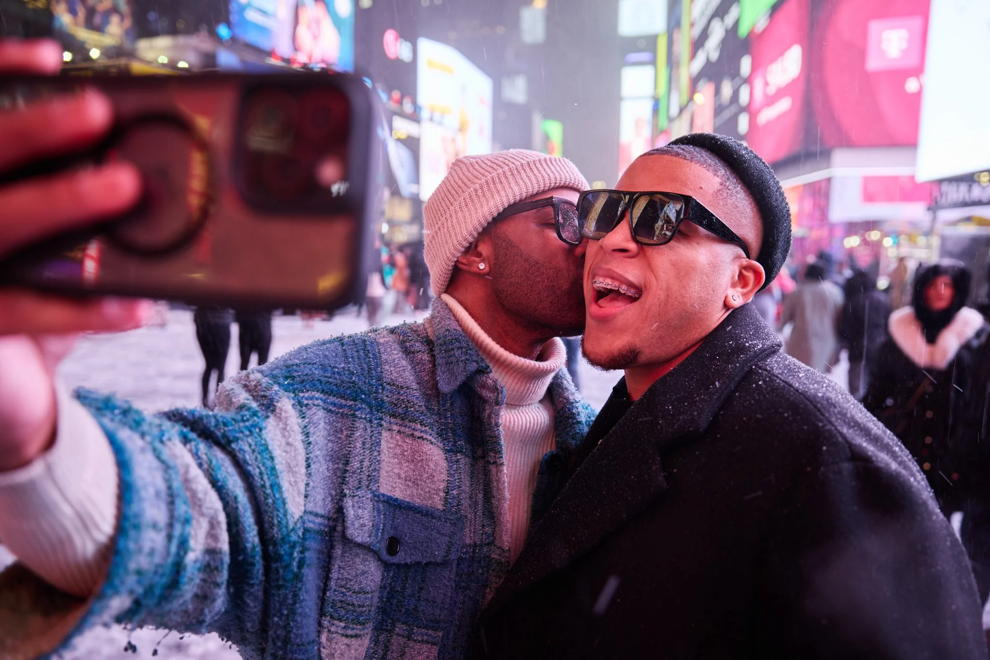 Two men are taking a selfie together in Times Square at night, with bright, colorful billboards and other people visible in the background. One man is leaning in to kiss the other on the cheek.