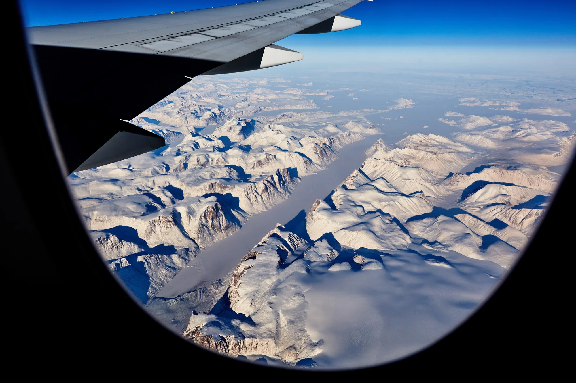 Aerial view of snow-covered mountains and a glacier at Auyuittuq National Park, Baffin Island, Nunavut, Canada, seen from an airplane window.