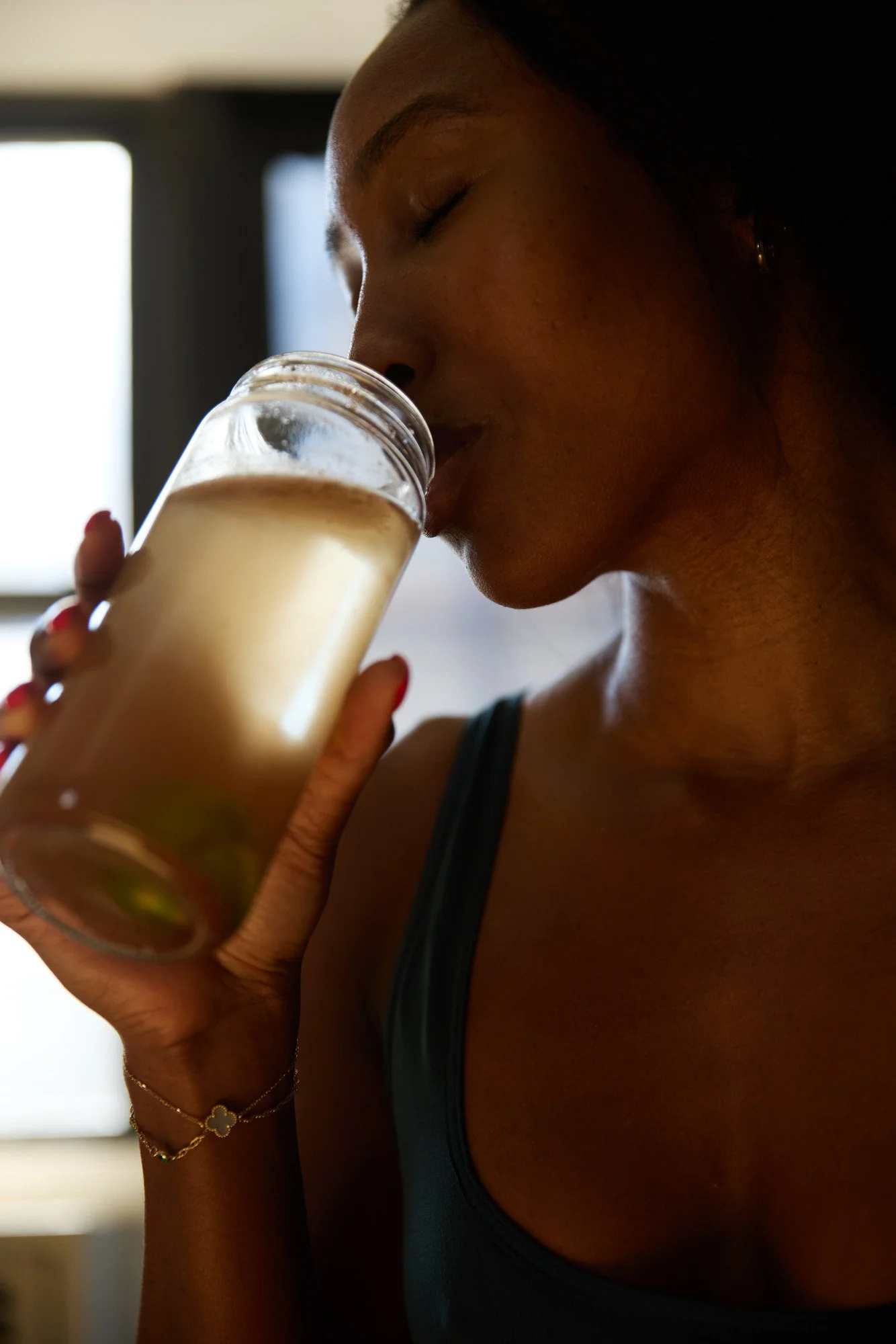 A young Black woman Francia Cooper with dark hair and earrings drinking a beige-colored beverage from a glass, wearing a dark sleeveless top, with a blurred background.