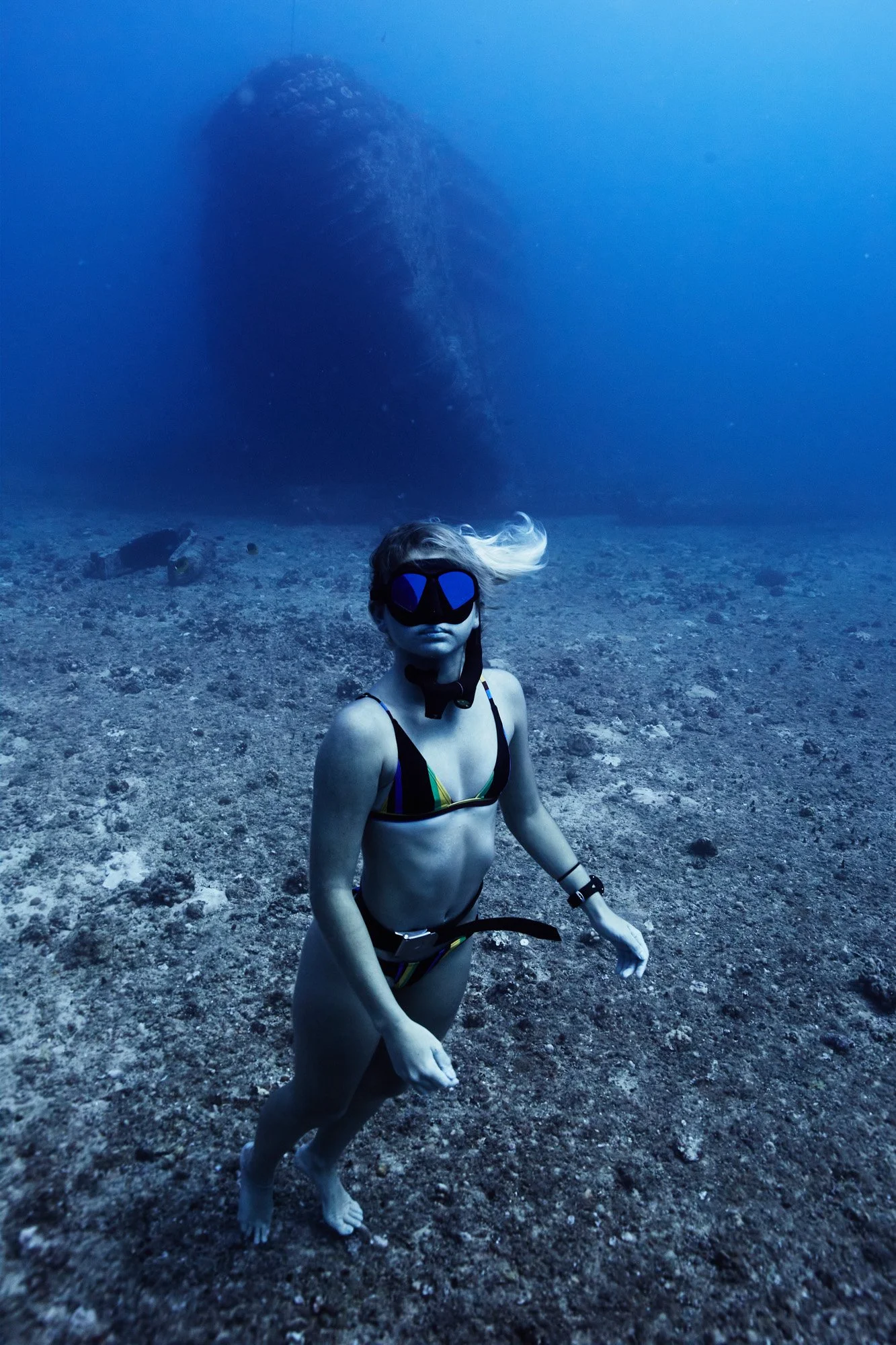 A woman with blonde hair, wearing a bikini, underwater in the ocean with a scuba mask, standing on the seabed near a large submerged shipwreck, Sea Tiger, off of Waikiki, Oahu, Hawaii