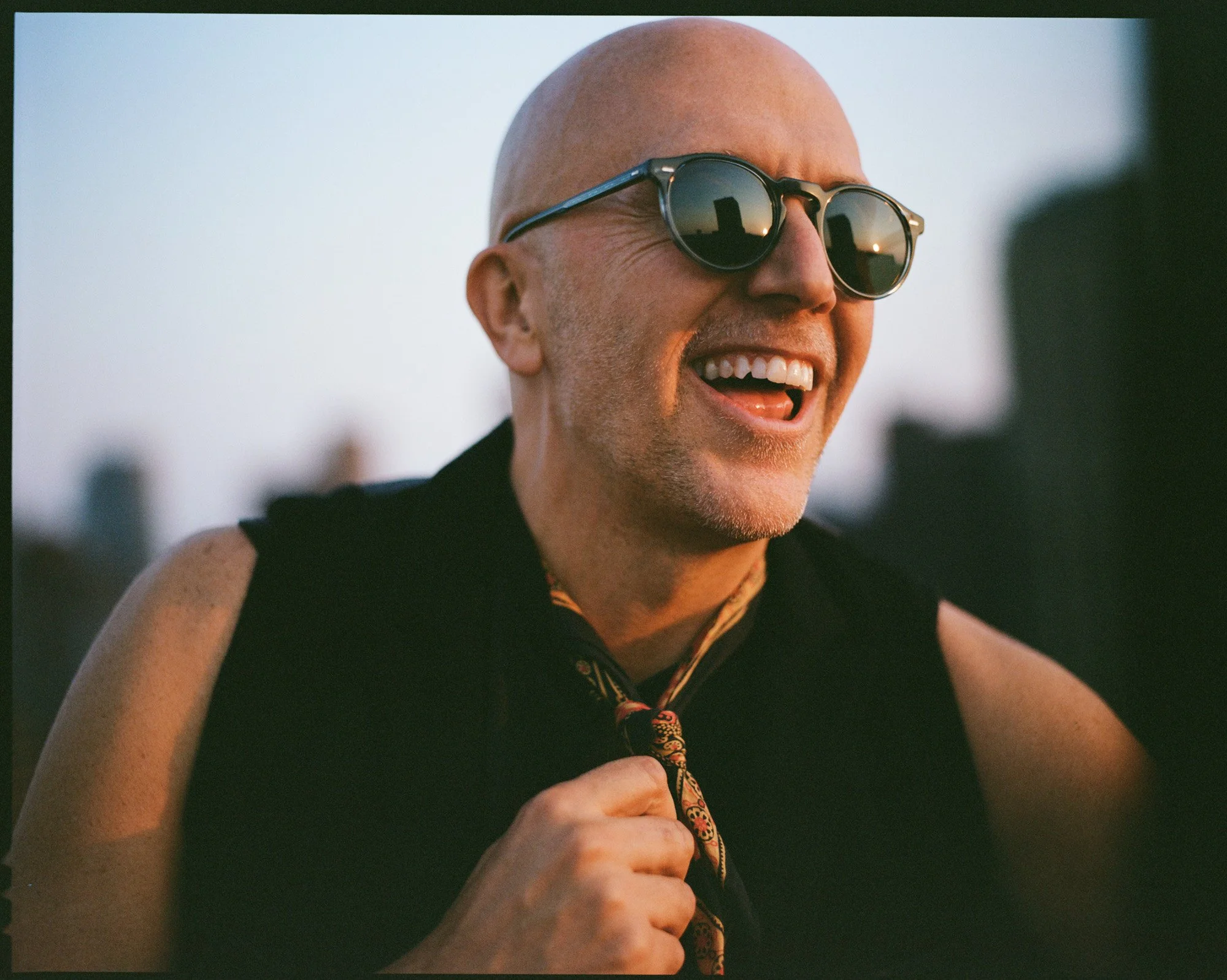 Lee Burridge smiling wearing sunglasses and a sleeveless black top, outdoors at sunset with the New York City skyline blurred in the background