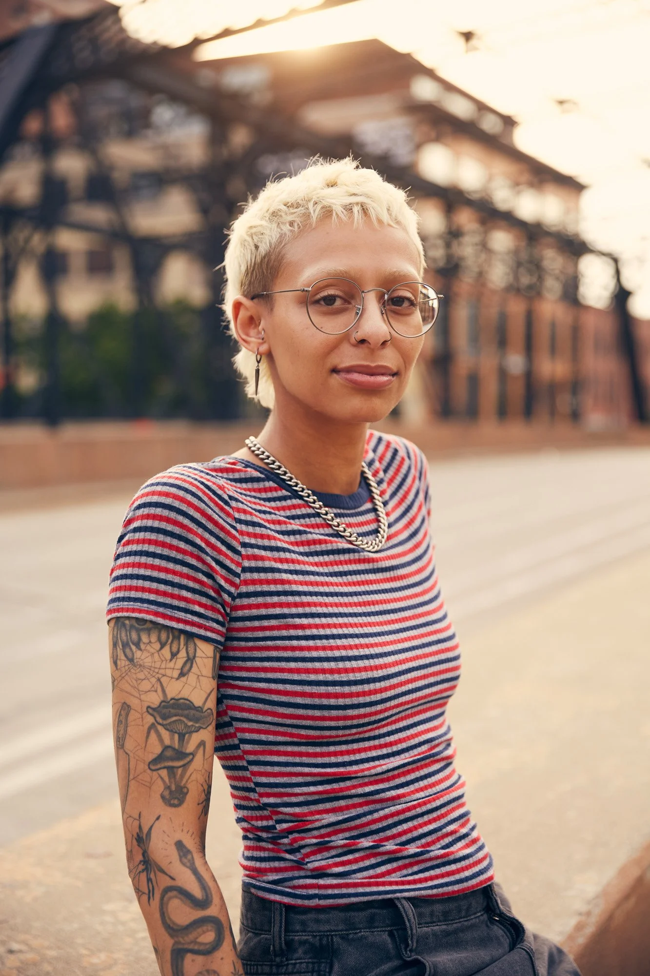 A woman with short blonde hair, glasses, tattoos on her arm, wearing a striped t-shirt, silver chain necklace, and earrings, standing outdoors on a city street during sunset in Minneapolis