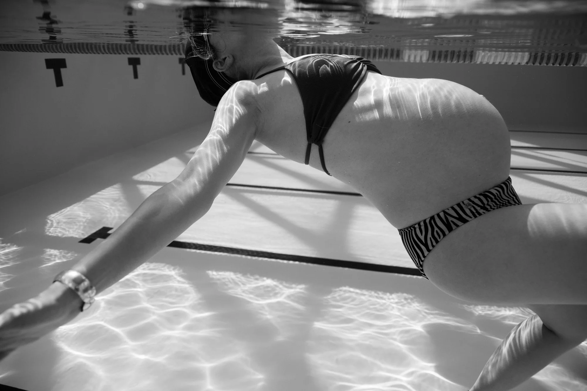 A pregnant woman in a swimsuit underwater, holding onto the pool's edge, with her hair partially floating and wearing a watch.