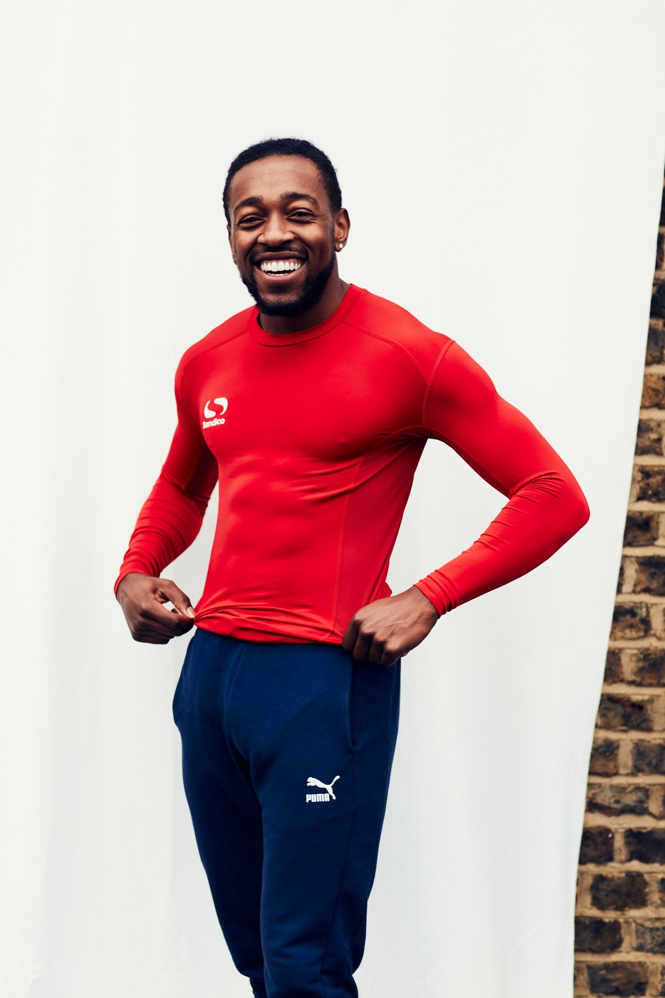 A smiling Black man wearing a red athletic top and navy blue Puma sweatpants standing in front of a plain white background with exposed brick wall on the right.