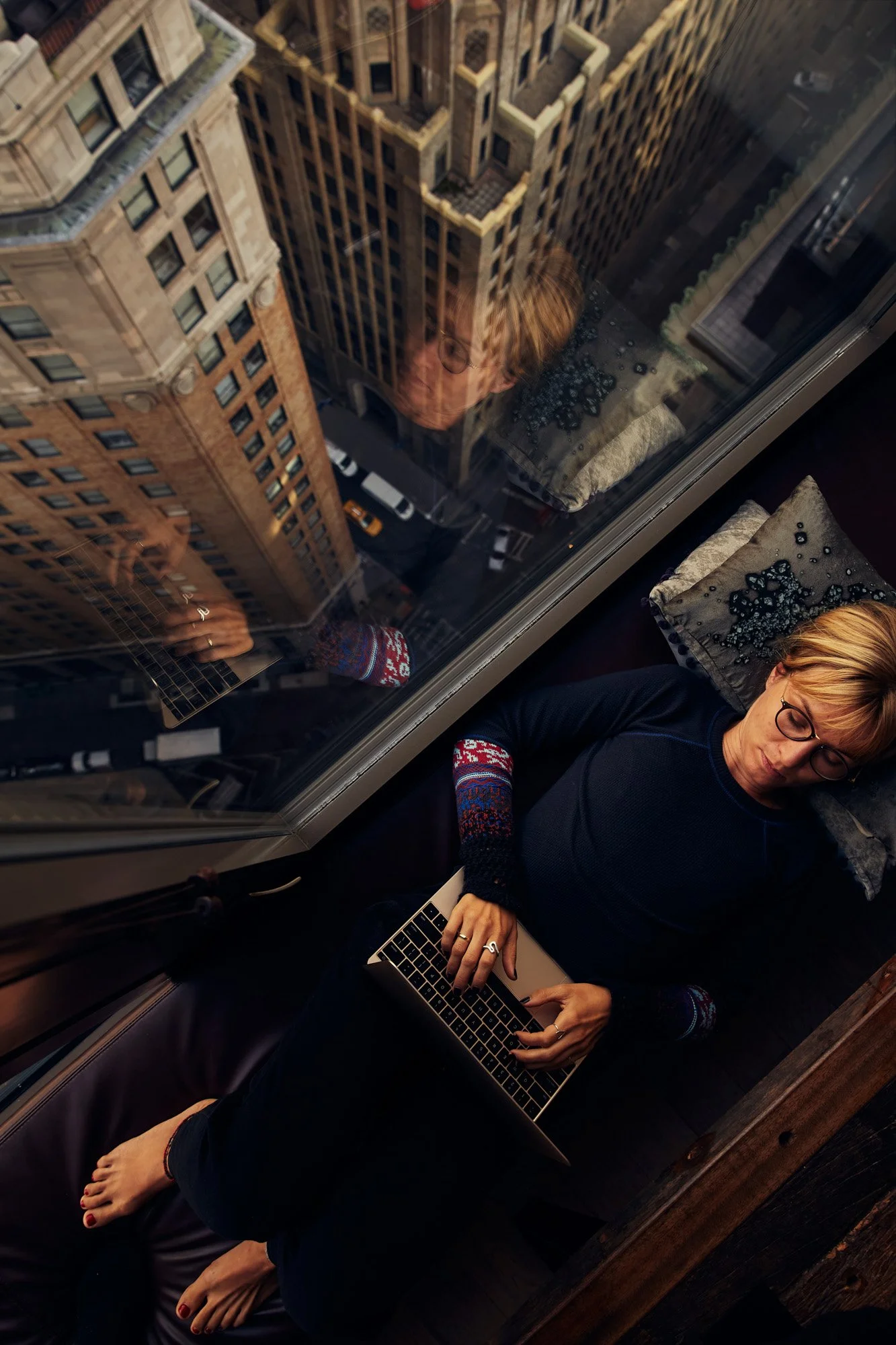 A woman with glasses, blonde hair, and a dark sweater with colorful patterned sleeves is lying on a bed next to a large window in a high-rise building in downtown Manhattan New York City, using a laptop. She has her eyes closed and seems relaxed.