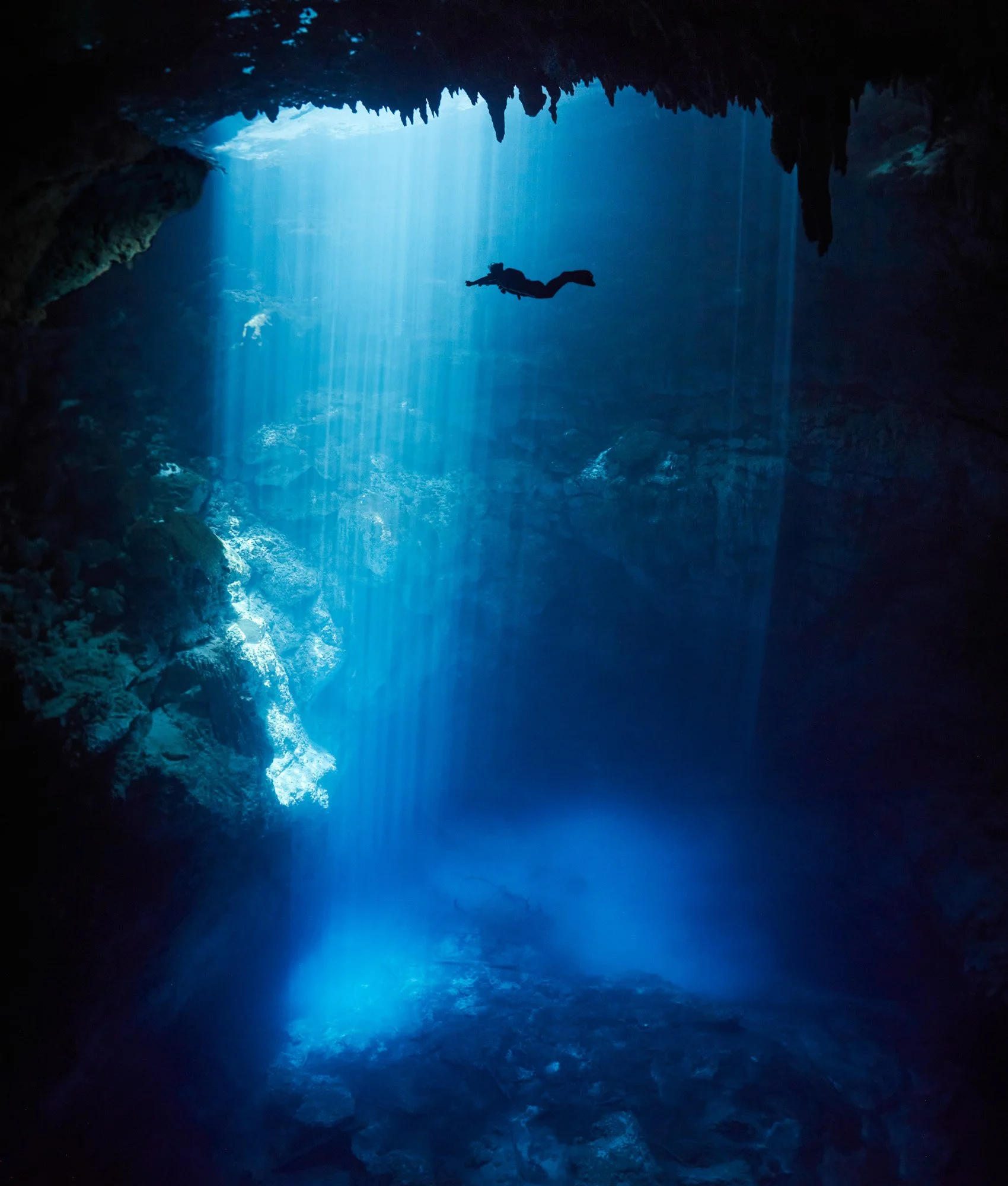 A scuba diver swimming in an underwater cave illuminated by sunlight streaming through the entrance of the cenote El Pit, near Playa del Carmen, in the Yucatan Peninsula of Mexico.