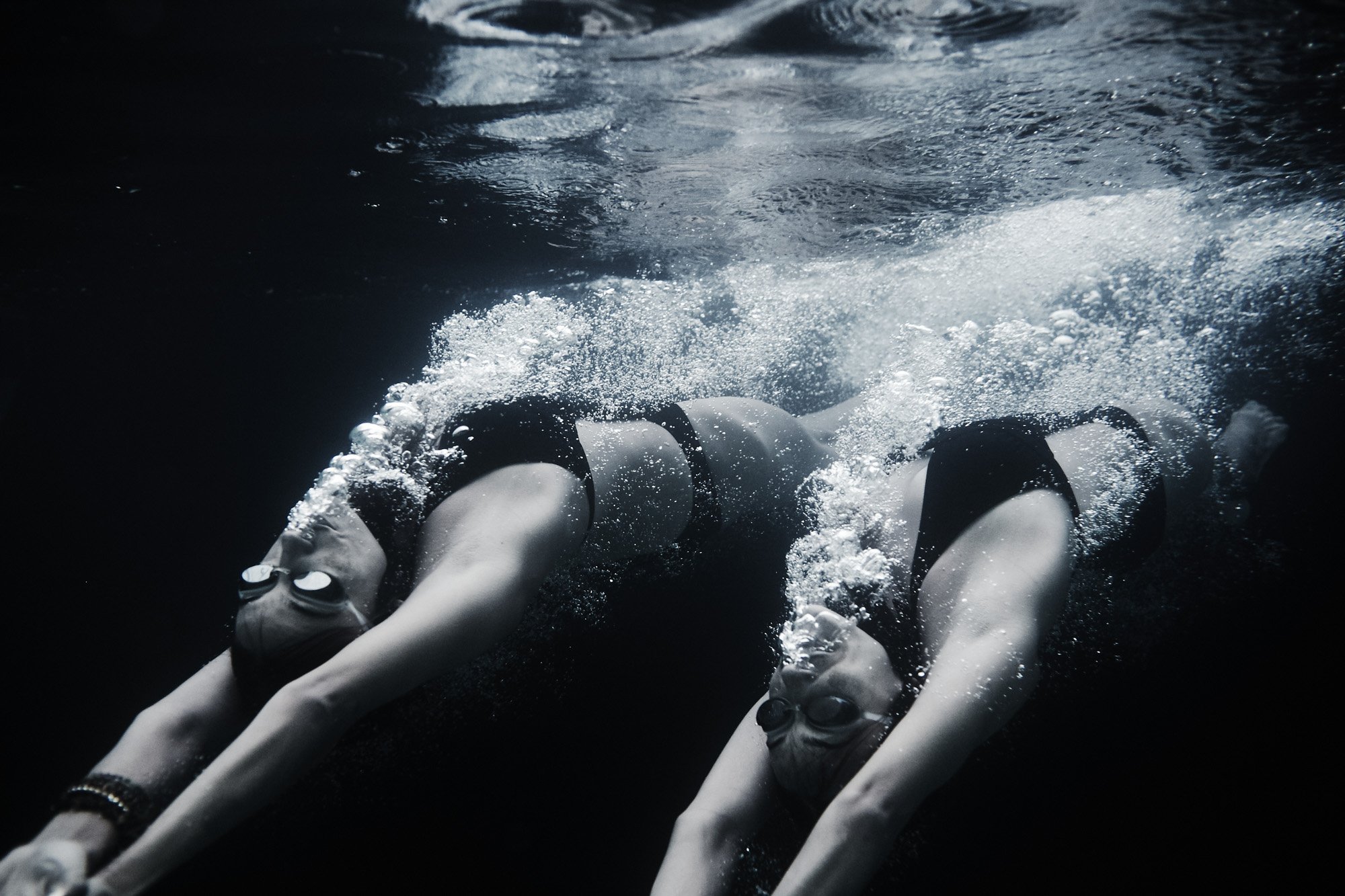 Two women swimming underwater wearing a black one-piece swimsuit and goggles, extending her arms forward, with bubbles trailing behind her.