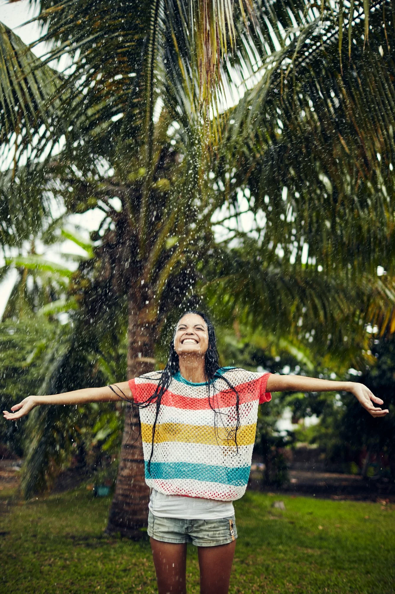 Young woman in Tahiti joyfully standing outdoors in a tropical environment with arms outstretched, smiling as water splashes around her, with a palm tree in the background.