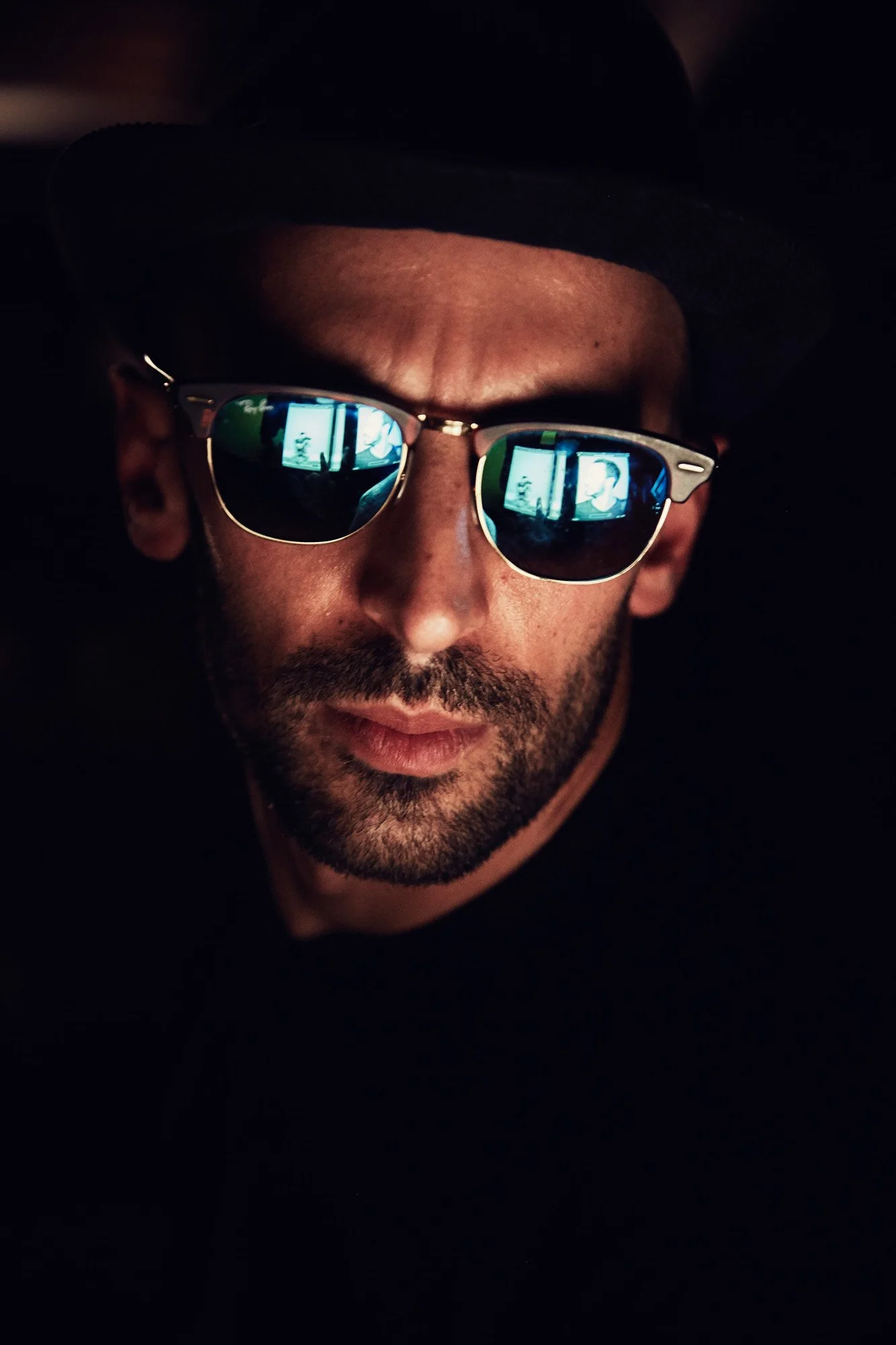 Close-up of artist JR wearing sunglasses, a hat, and a dark shirt, with reflection of screens in his sunglasses.