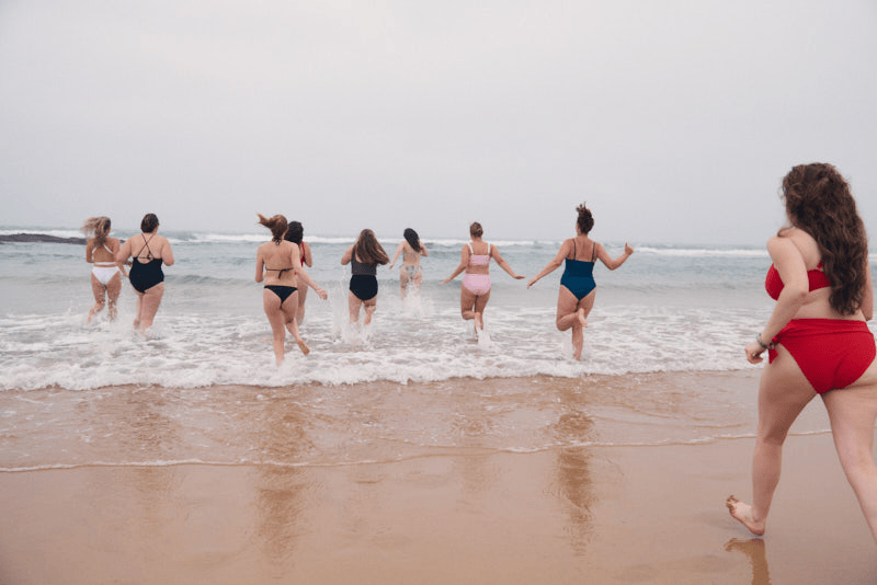 Group of women in swimsuits entering the ocean at the beach, with one woman running toward the rest of the group and gives her daughter a hug.
