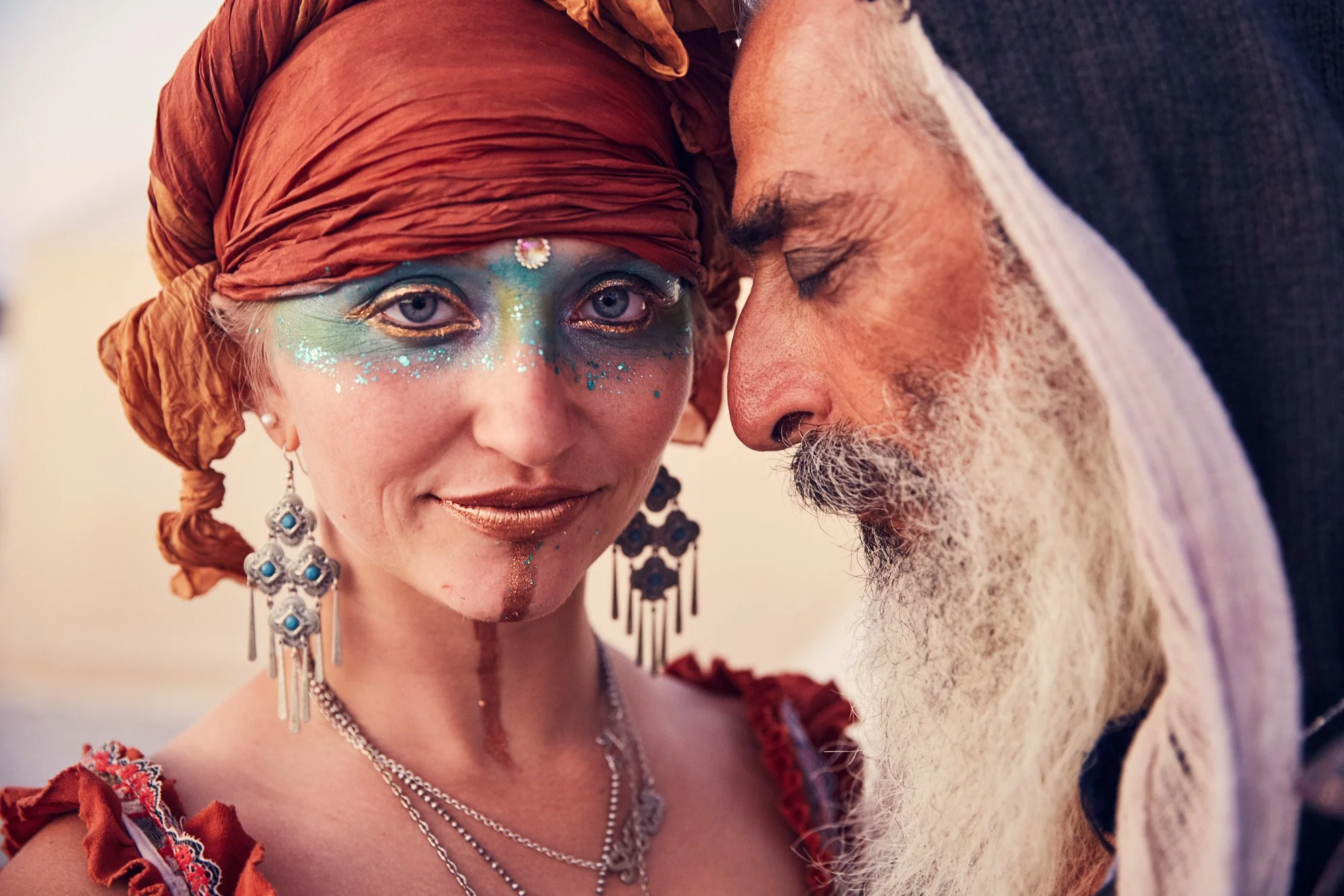 A woman with elaborate face paint, sparkles, and jewelry, wearing a brown headscarf, and an older man with a gray beard and mustache with his eyes closed, leaning their foreheads together. Burning Man