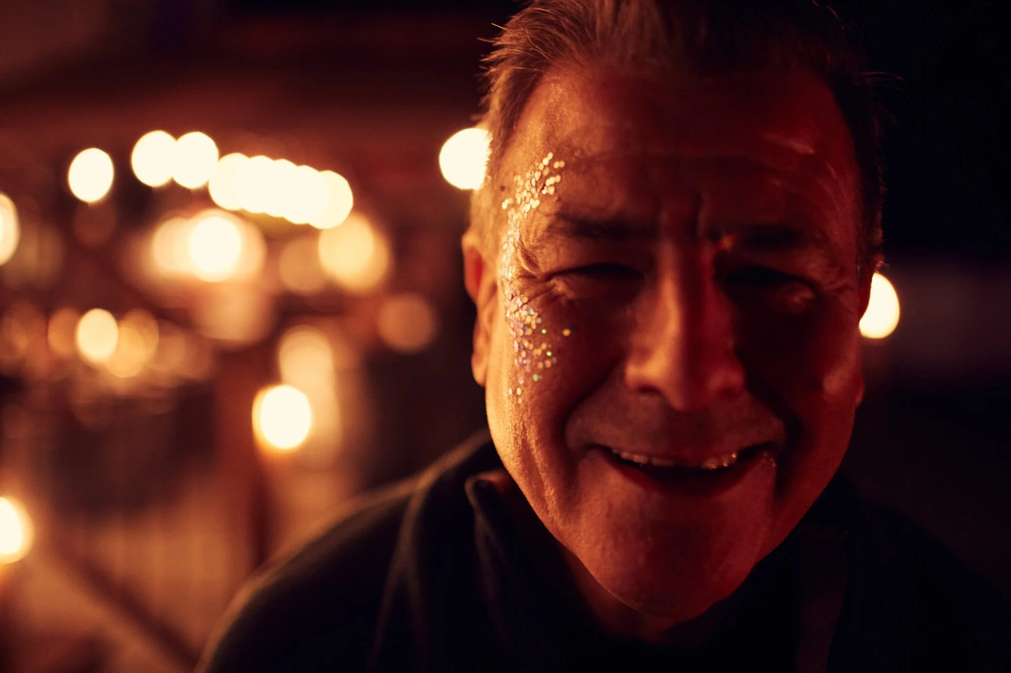 A person smiling in a dimly lit setting with warm, blurry string lights in the background, some glitter or sparkles on his face.