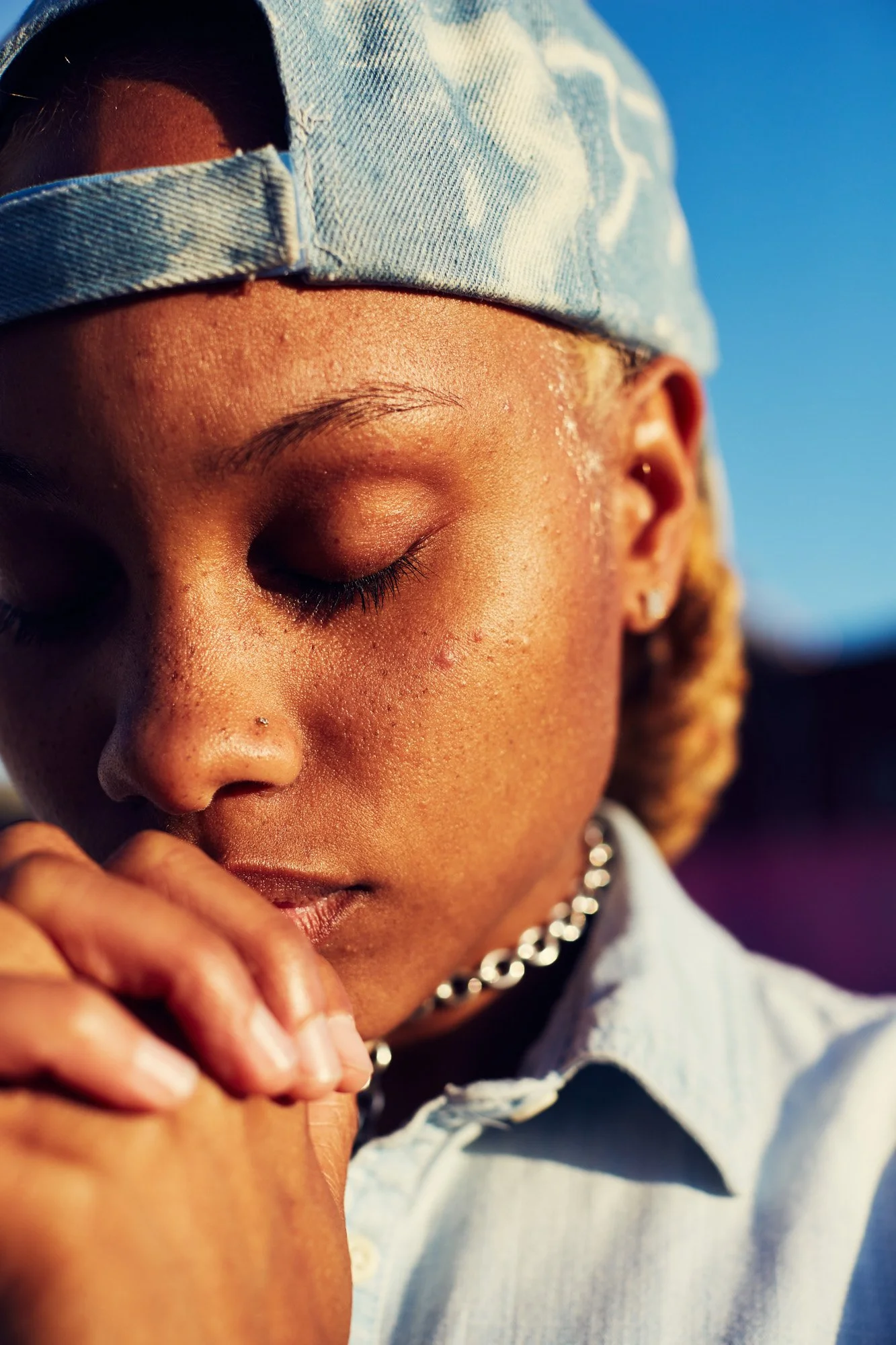 A person with closed eyes, wearing a light blue denim cap, a white shirt, and a silver chain, appears to be in a moment of reflection or prayer, with their hand resting on their face.