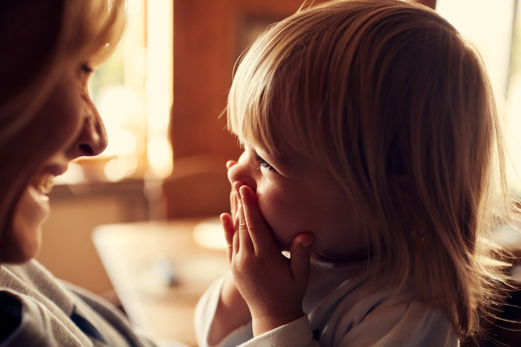 A young child with blonde hair covers their mouth with her hands as she laughs while looking at an adult woman, who is smiling at her. The scene is warmly lit with soft background lighting.