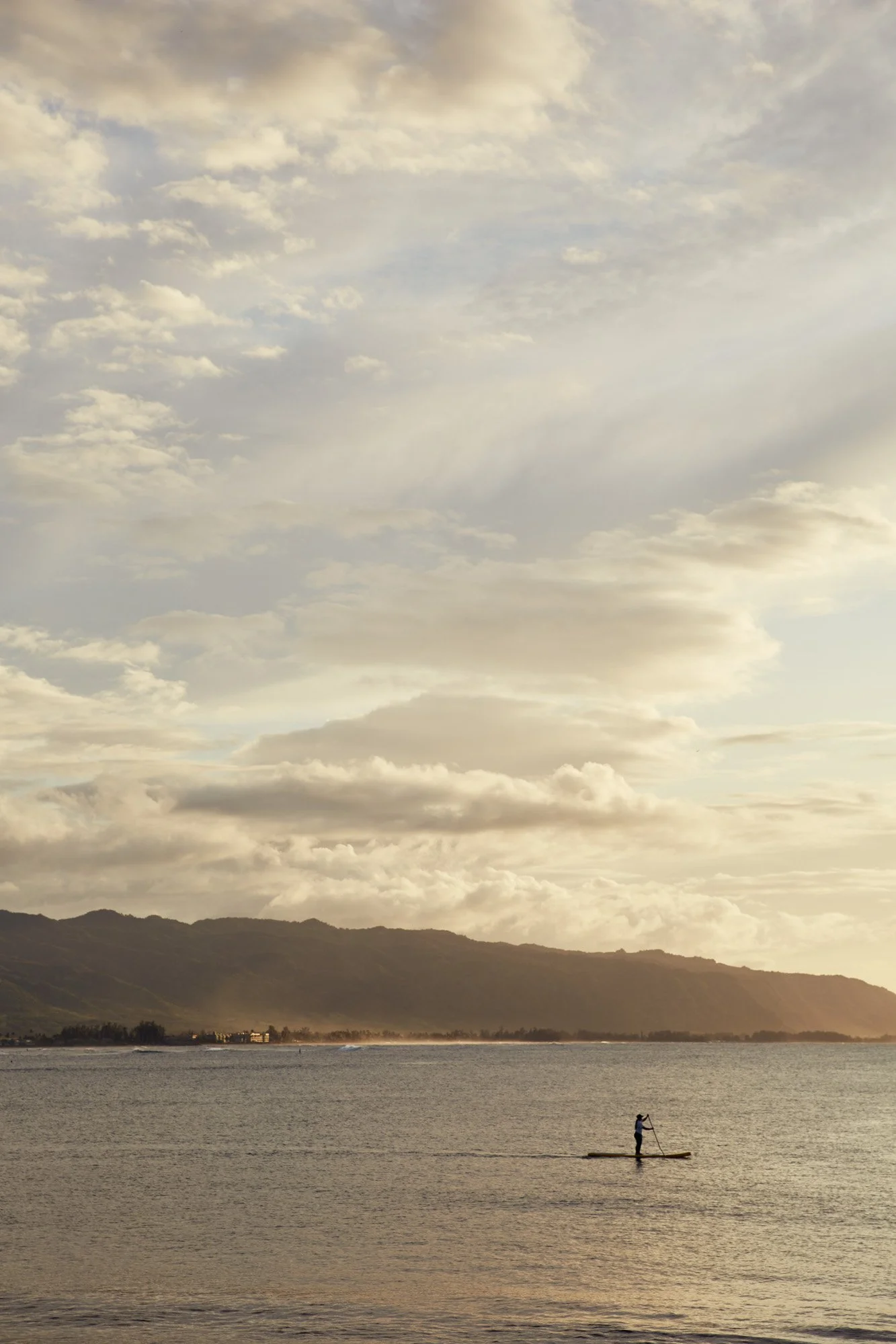 A person paddleboarding on the water near a mountainous coastline at sunset or sunrise, with a partly cloudy sky overhead. Haleiwa, North shore Oahu Hawaii