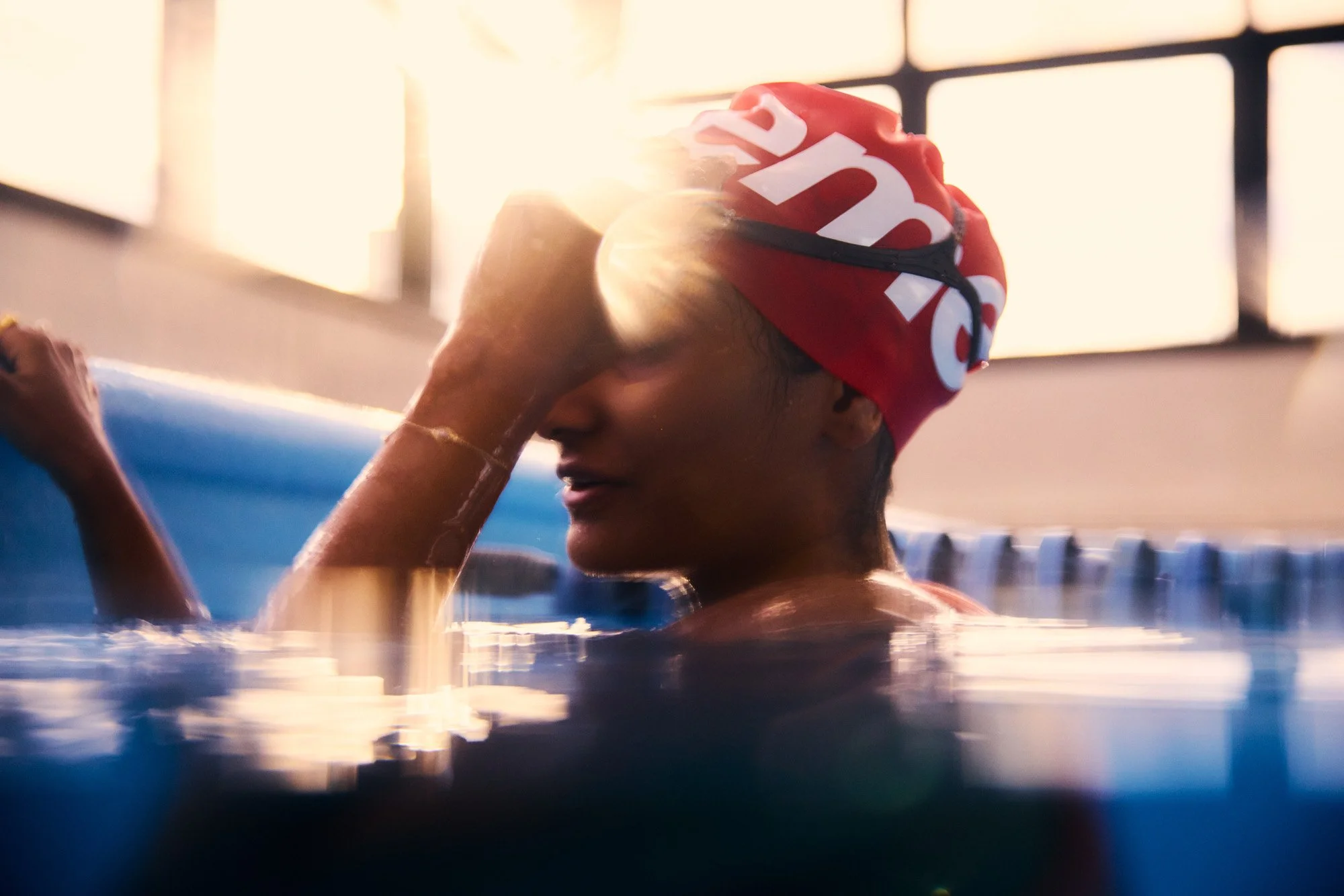 A swimmer wearing a red swim cap with white writing is in a pool, adjusting goggles with sunlight coming through large windows behind.