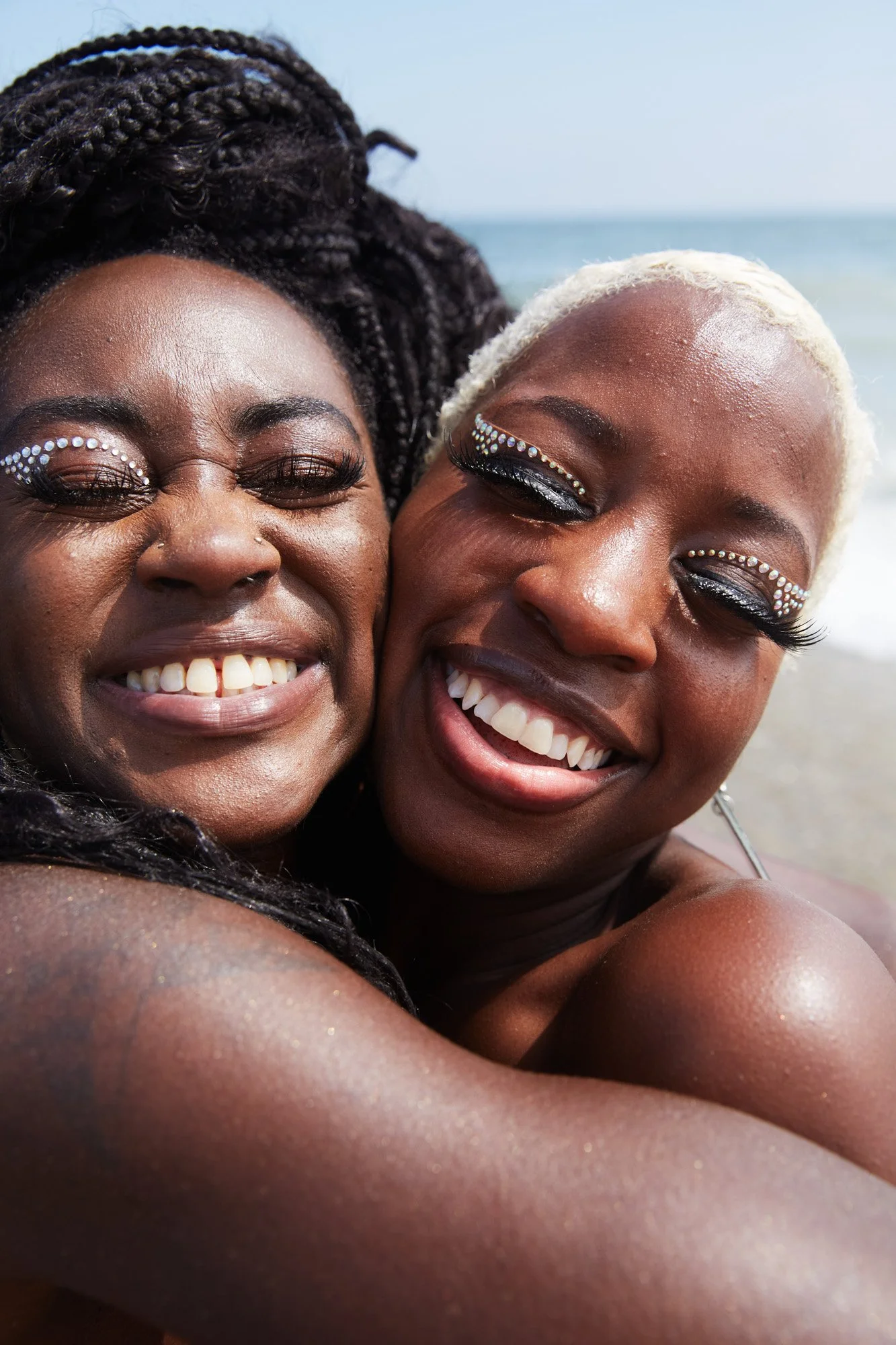 Two women smiling and hugging at the beach, with ocean in the background. They have decorative eye makeup with rhinestones. Pride Jacob Riis Beach New York City