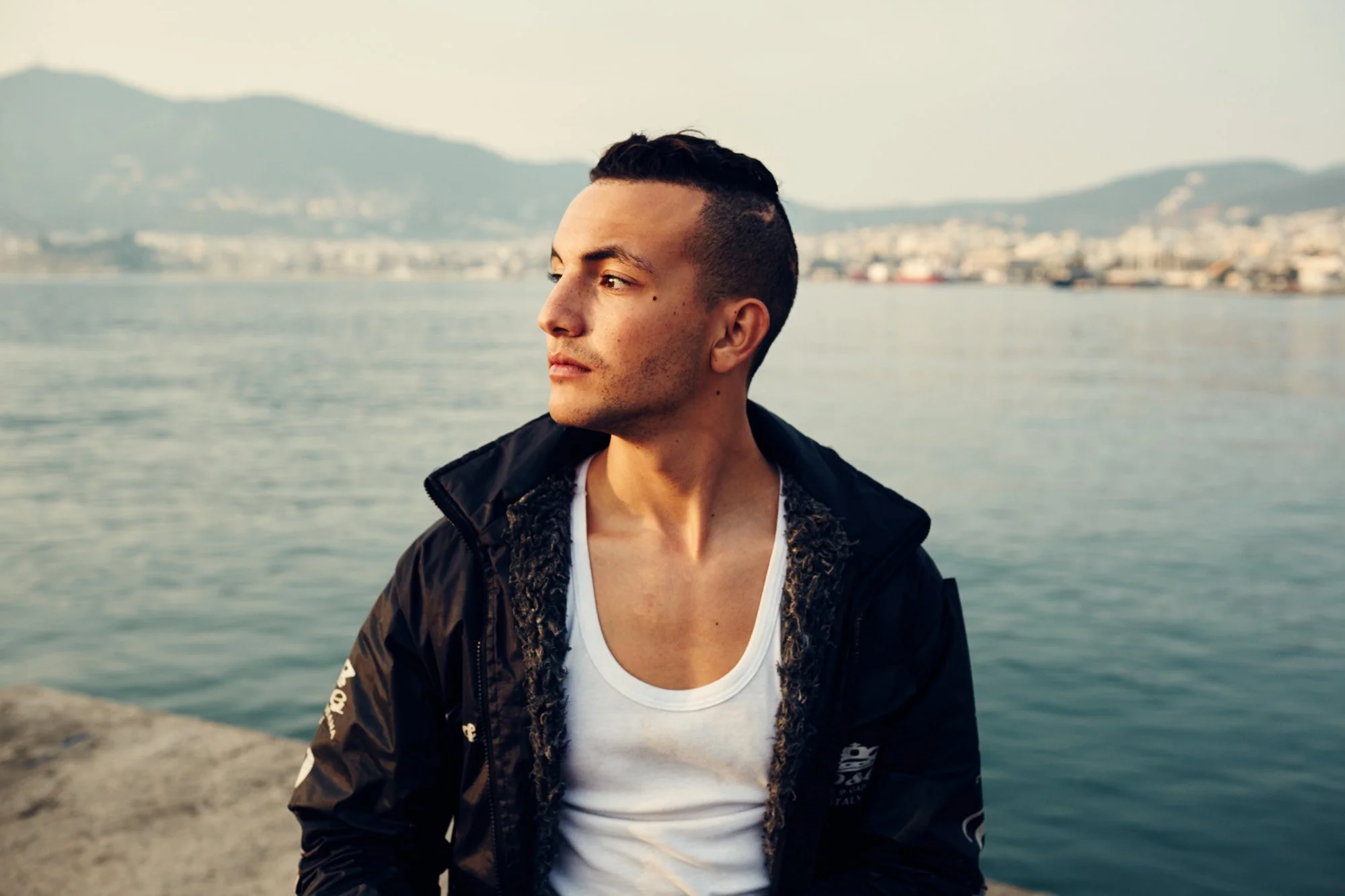 A young man with short dark hair and light skin looking to the side, wearing a black jacket over a white shirt, sitting near a body of water with a cityscape and mountains in the background during the daytime. Lesbos Greece