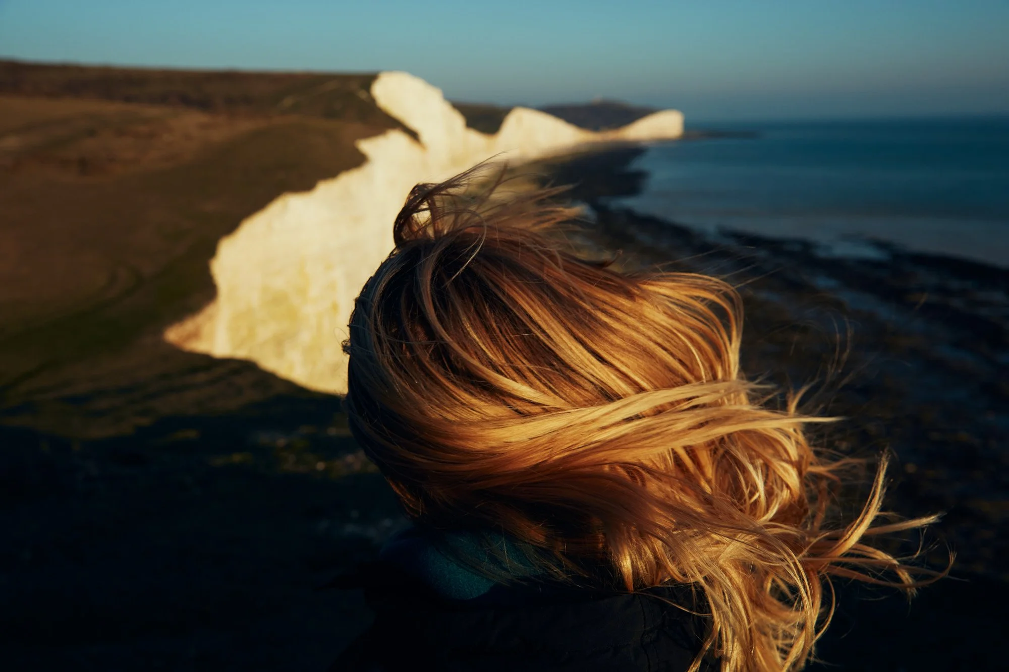 Young woman with blonde hair standing on a cliff overlooking the ocean at sunset, with white chalk cliffs of Seven Sisters England in the background