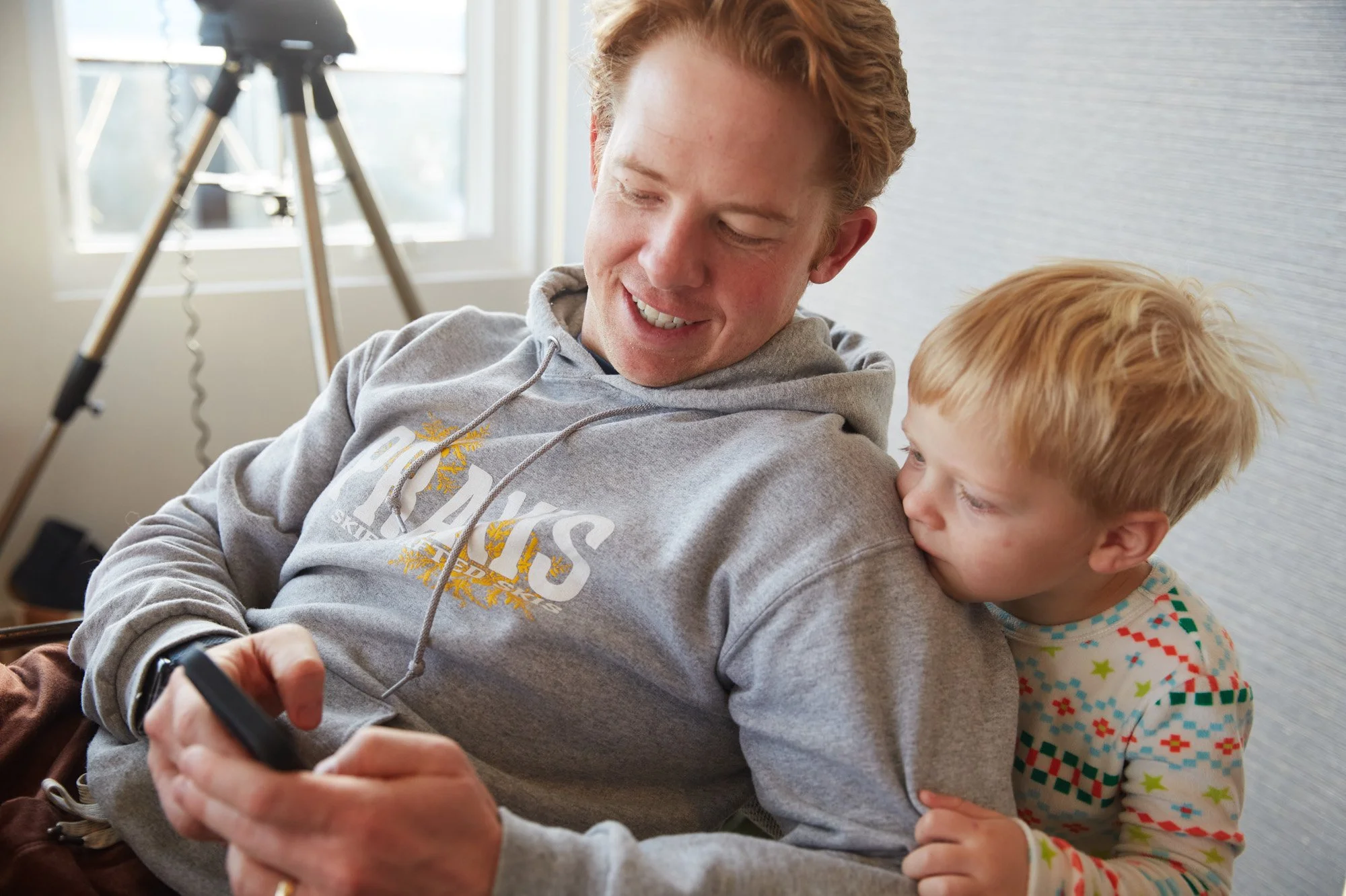 A young man with curly hair and a gray hoodie smiling while looking at his phone, with a young boy with light hair hugging him from behind and looking at his shoulder, in a bright room with a camera on a tripod in the background.