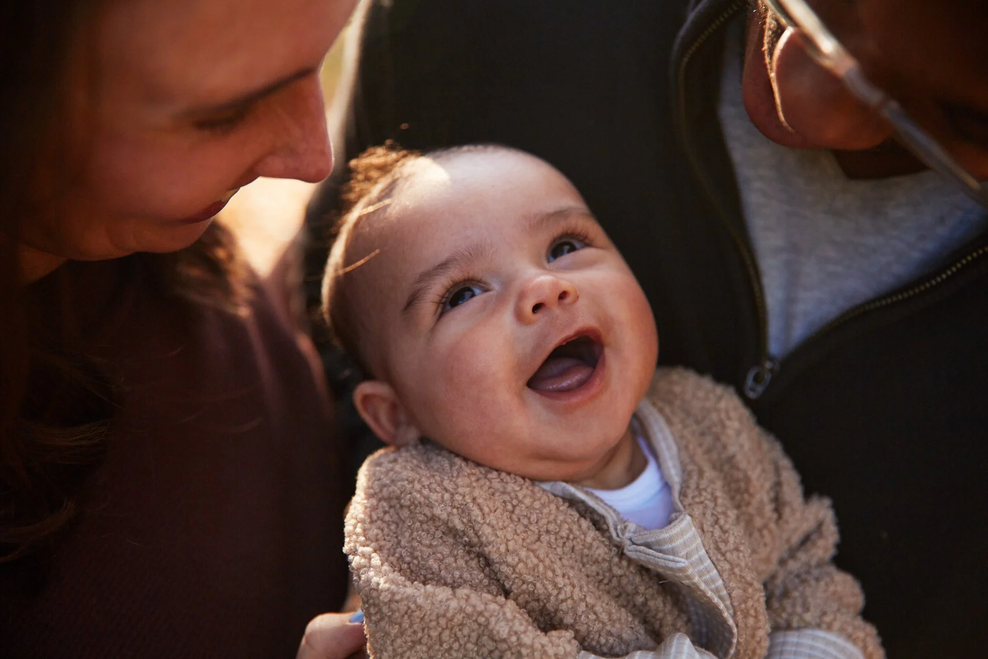 A smiling baby looking up at a woman and a man, likely her parents, in close-up. The baby has a joyful expression, showcasing a wide smile and bright eyes, with a fuzzy beige jacket.