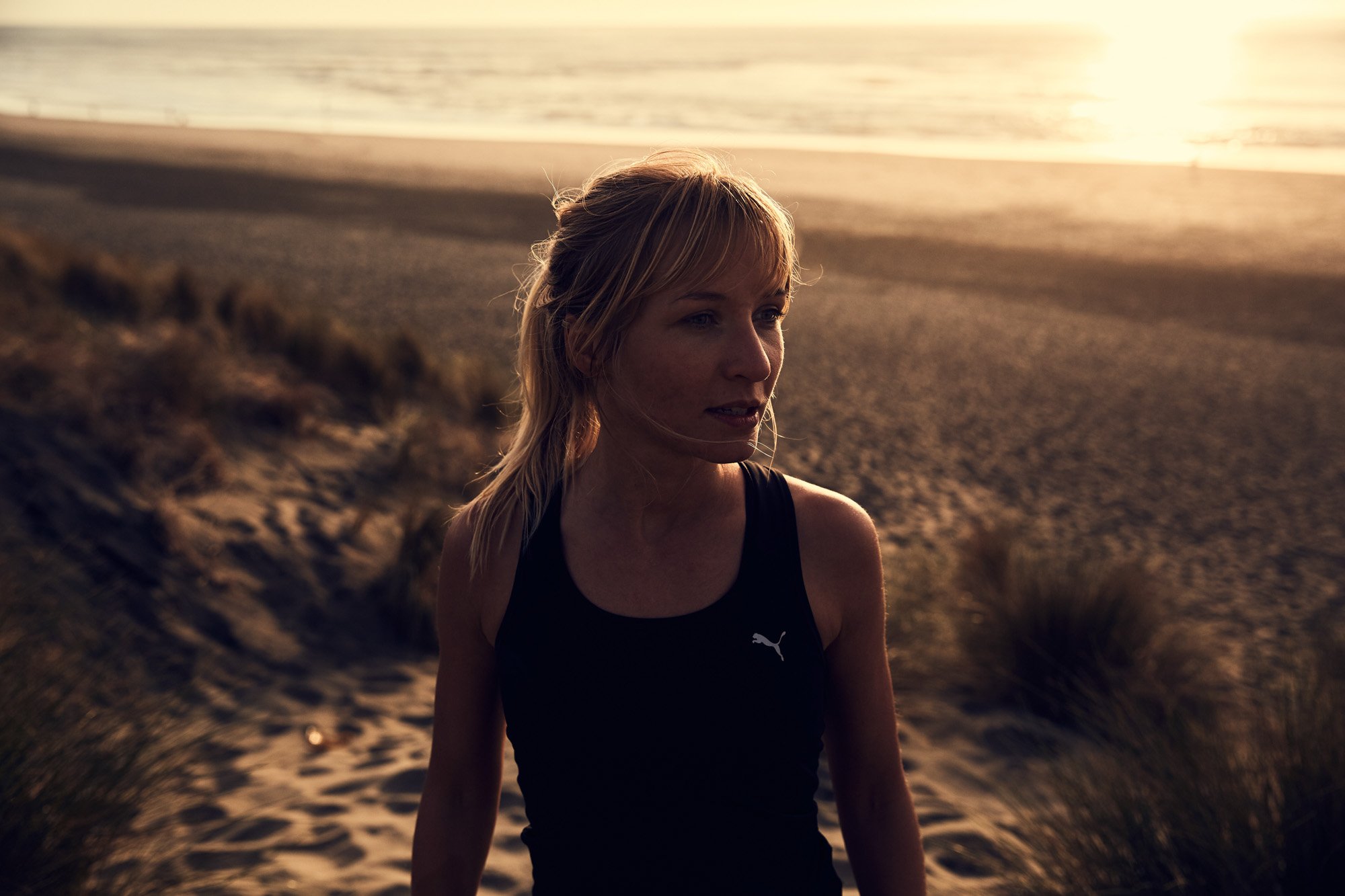 A woman with blonde hair in a ponytail wearing a black Puma tank top, standing outdoors at sunset on the dunes at Ocean Beach San Francisco with the ocean in the background.