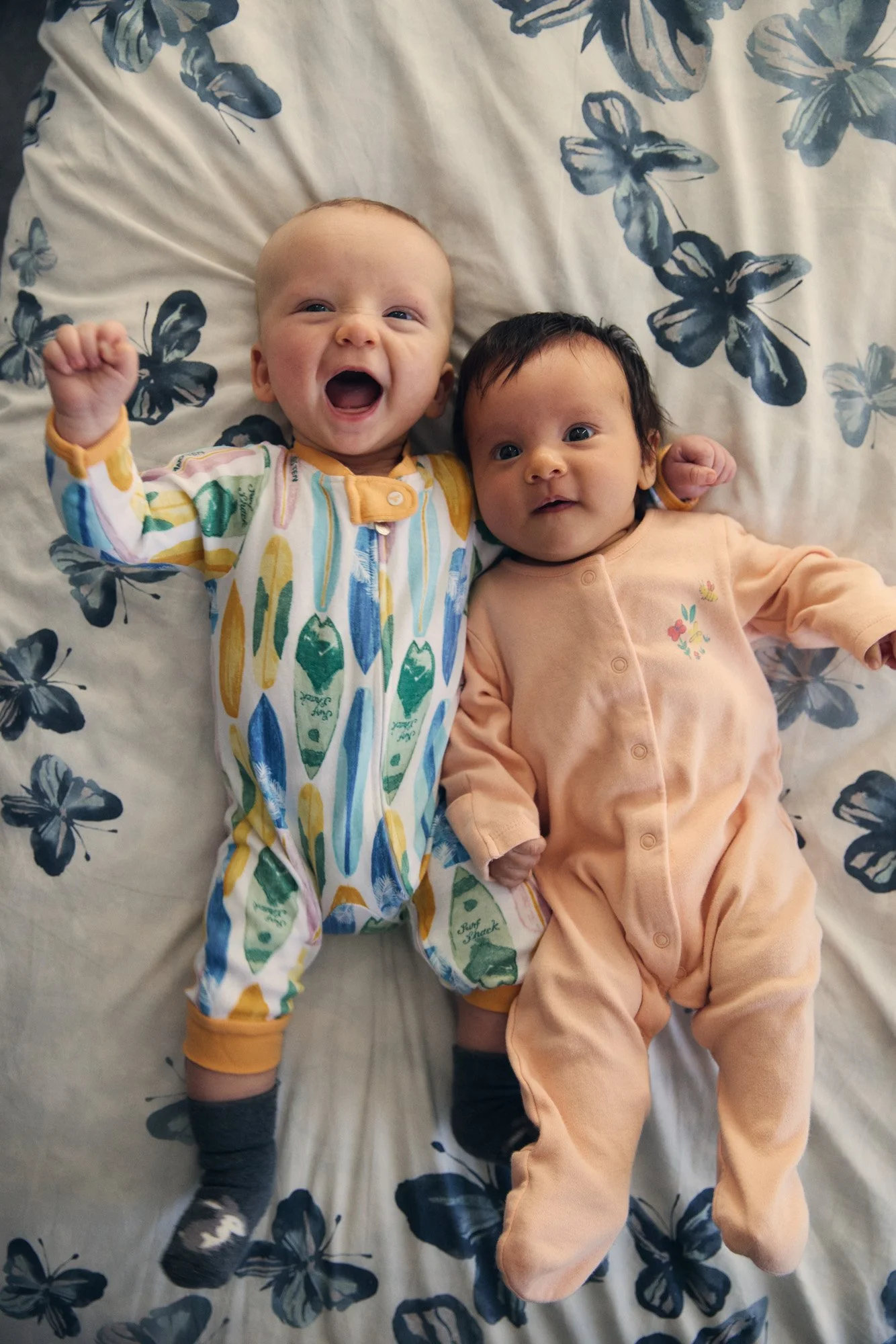 Two babies lying on a bed with butterfly-patterned sheets, smiling and one making a fist.
