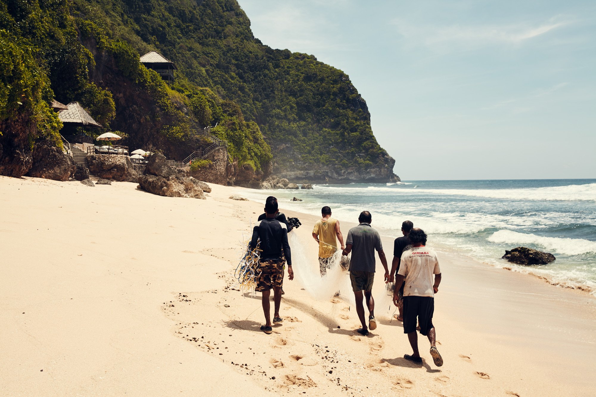 Group of five people walking along a sandy beach near the ocean with a green, rocky hillside in the background near the Bulgari Resort on Bali.