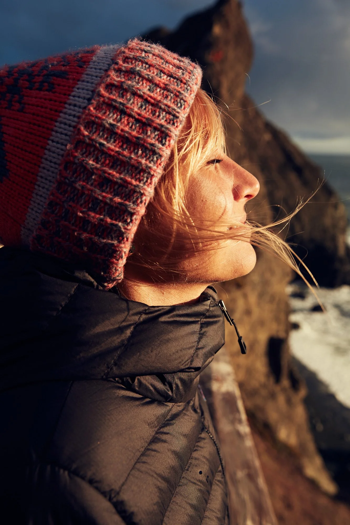 A woman with blonde hair looking towards the setting sun, wearing a colorful knit hat and a black puffer jacket, standing outdoors near a rocky coastline, on Point Bonita in Marin County California near San Francisco