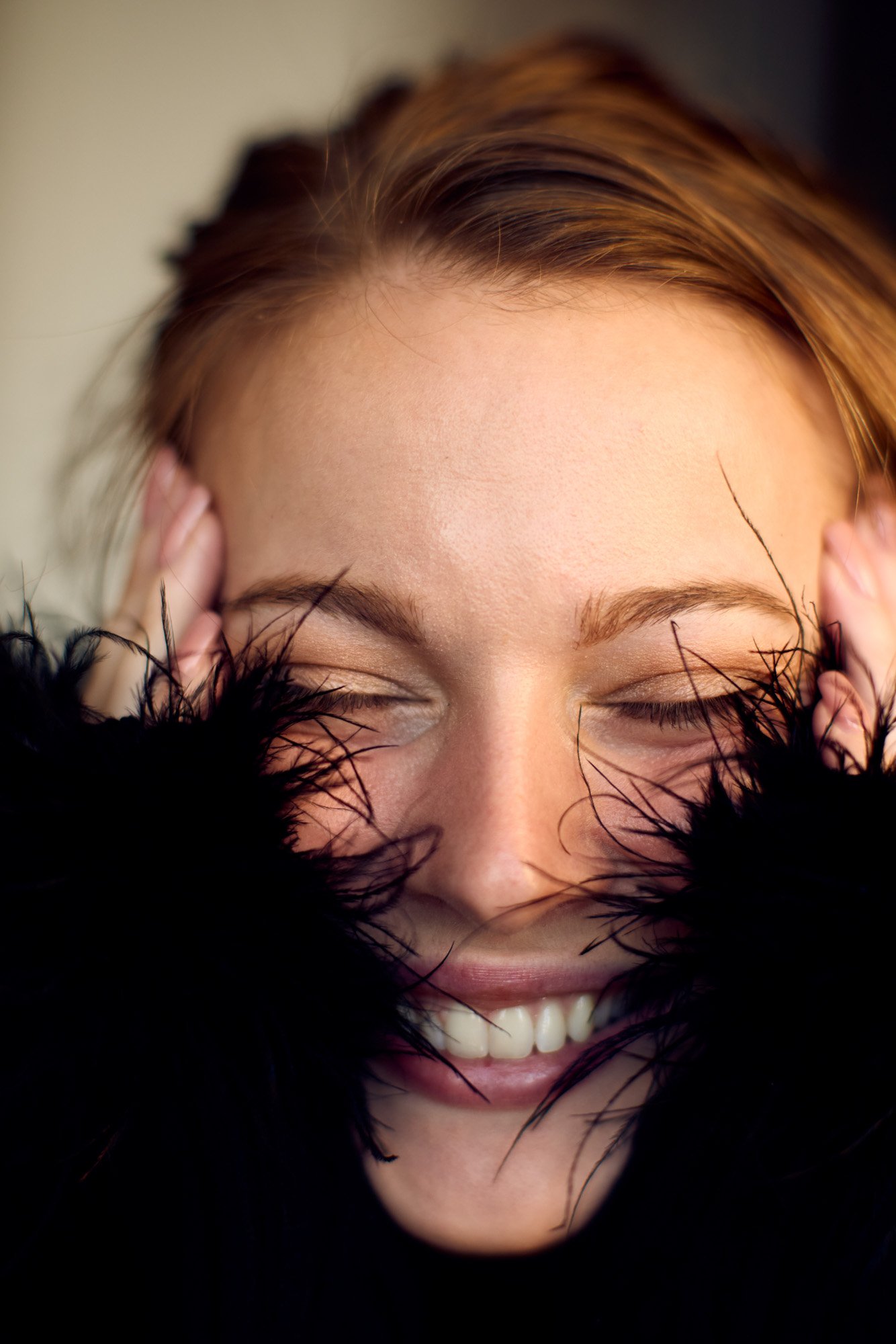 Close-up of a smiling young woman Taylore Crosley with closed eyes, wearing a black feathered garment, with her hands on her head, showcasing her hair and earrings.
