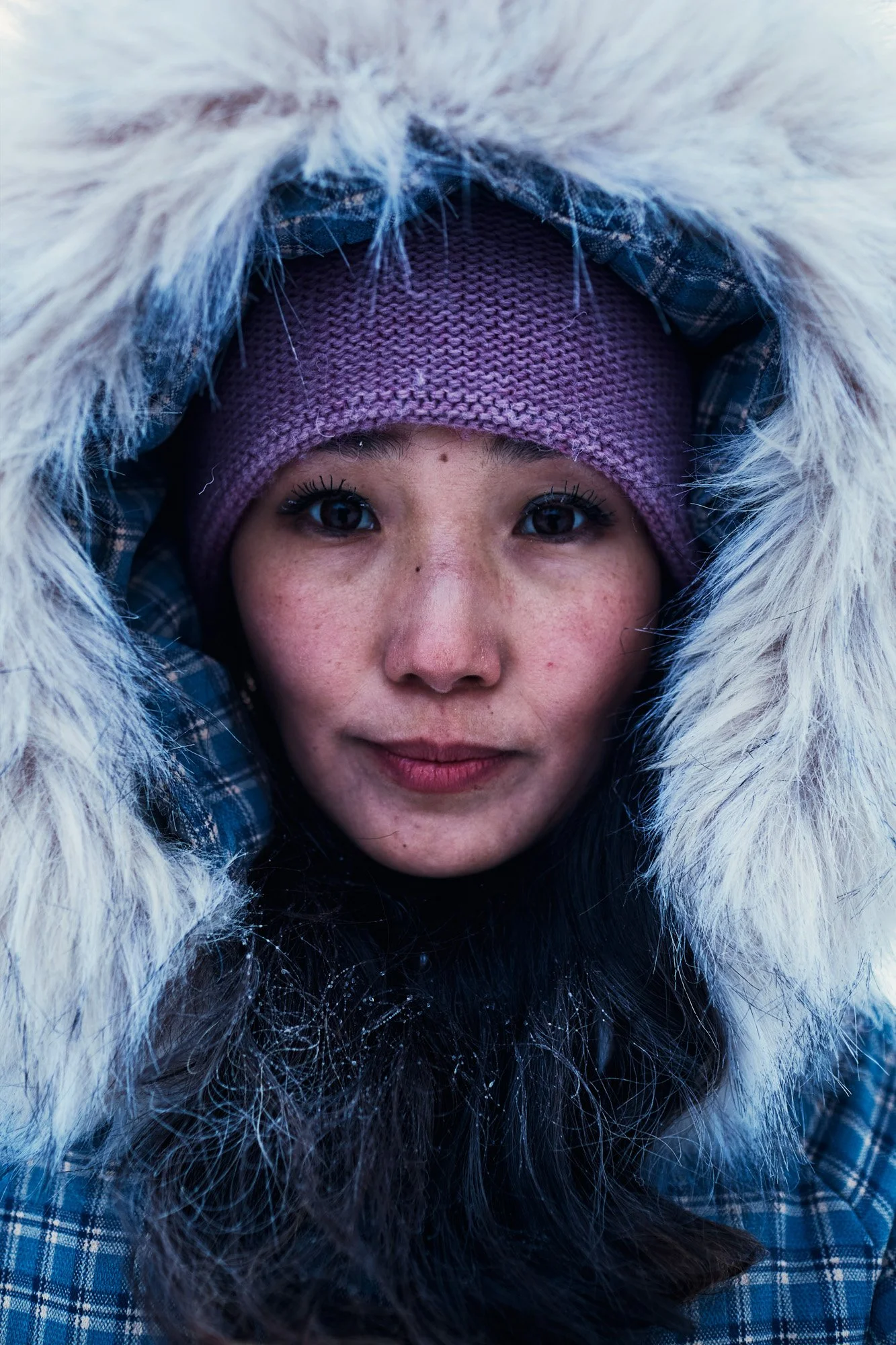 Close-up of a woman with a fair complexion and dark hair wearing a purple knit hat and a blue plaid coat with a fur-lined hood. Yakutsk Far East Siberia Russia